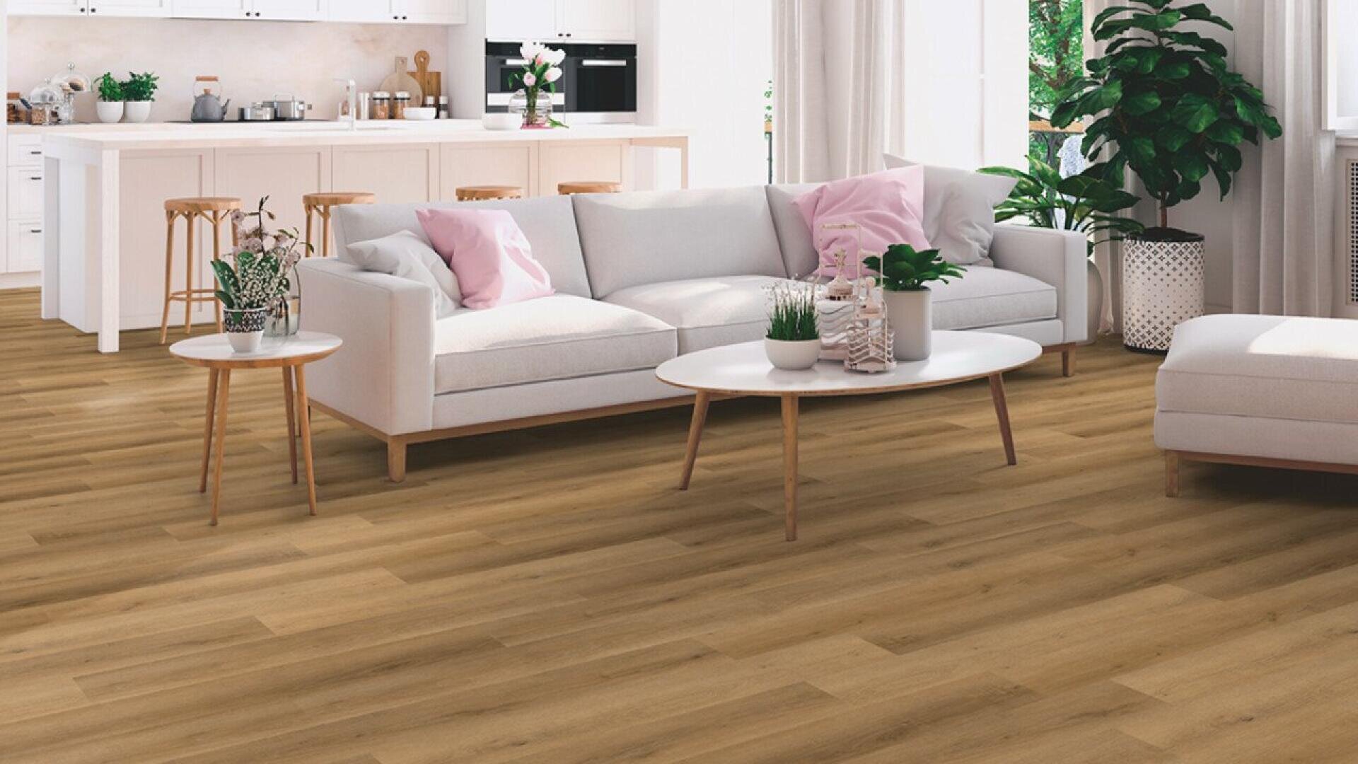 A modern living room with light wood flooring, a white sofa with pink and gray cushions, a round white coffee table, green plants, and a bright kitchen with bar stools in the background.