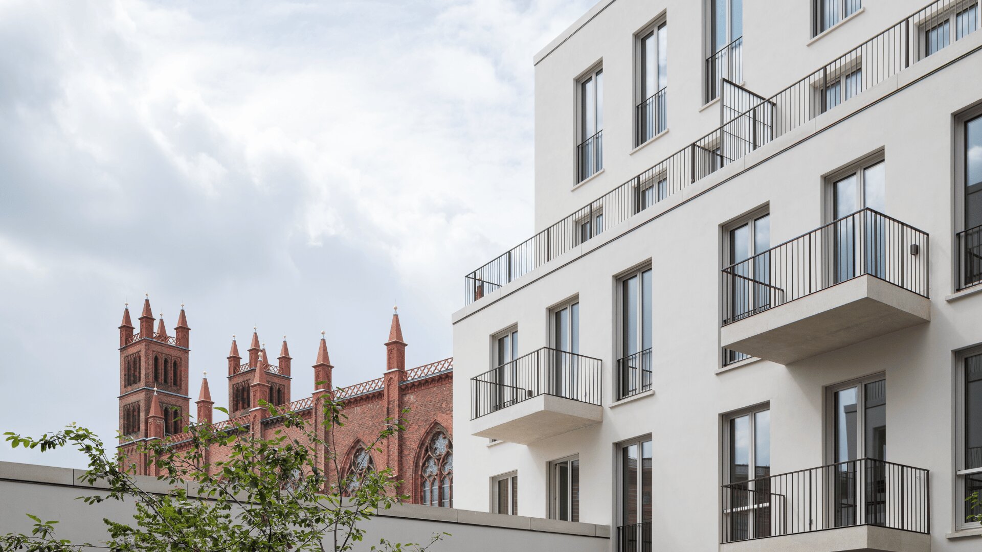 Im Vordergrund steht ein modernes weißes Wohnhaus mit Balkonen, während im Hintergrund unter einem bewölkten Himmel eine historische Kirche aus rotem Backstein mit hohen Türmen zu sehen ist.