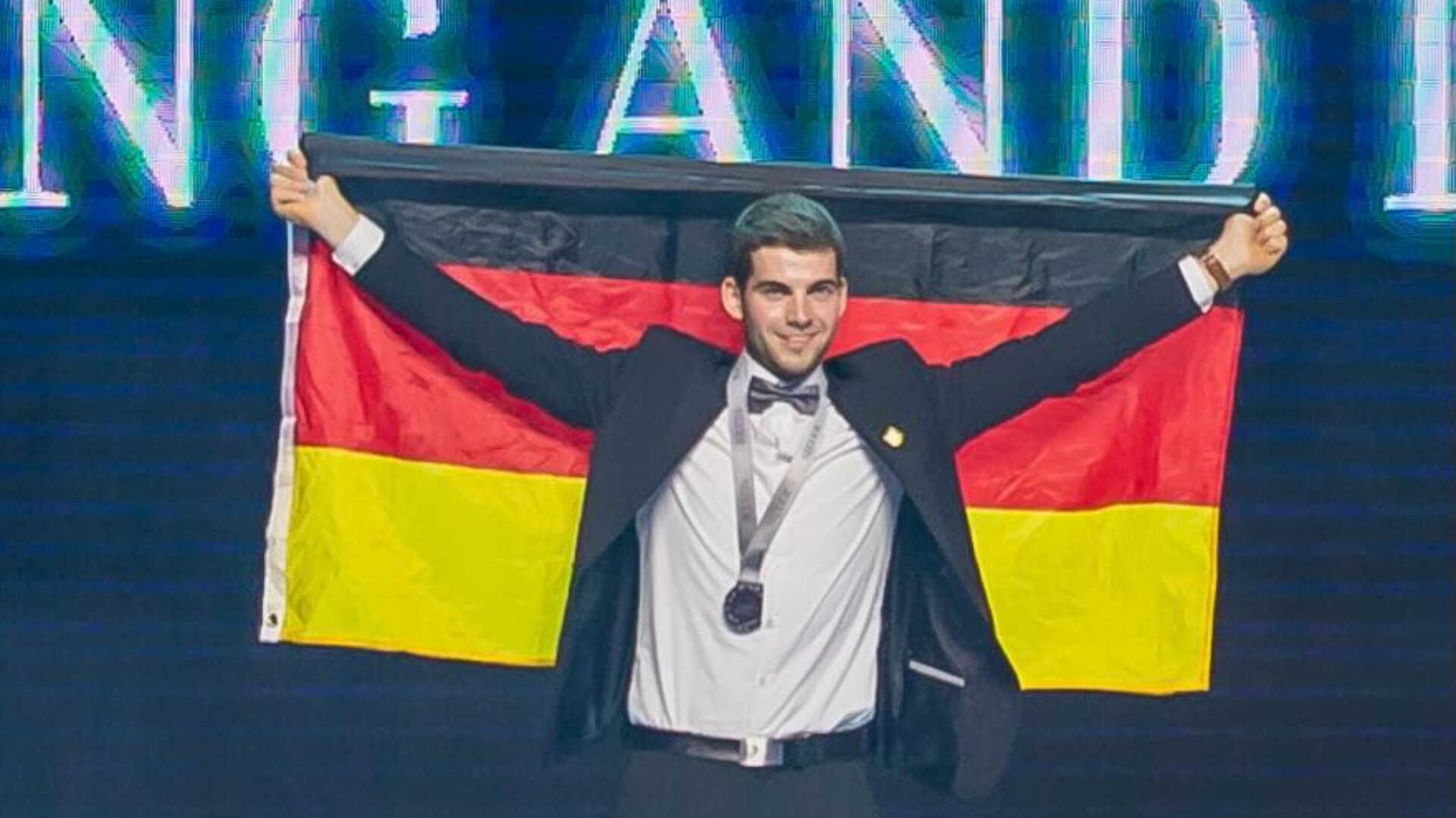 Eine Person in Anzug und Fliege hält eine deutsche Flagge hinter sich hoch und trägt eine Medaille um den Hals. Sie steht auf einer Bühne mit hellen Lichtern im Hintergrund.