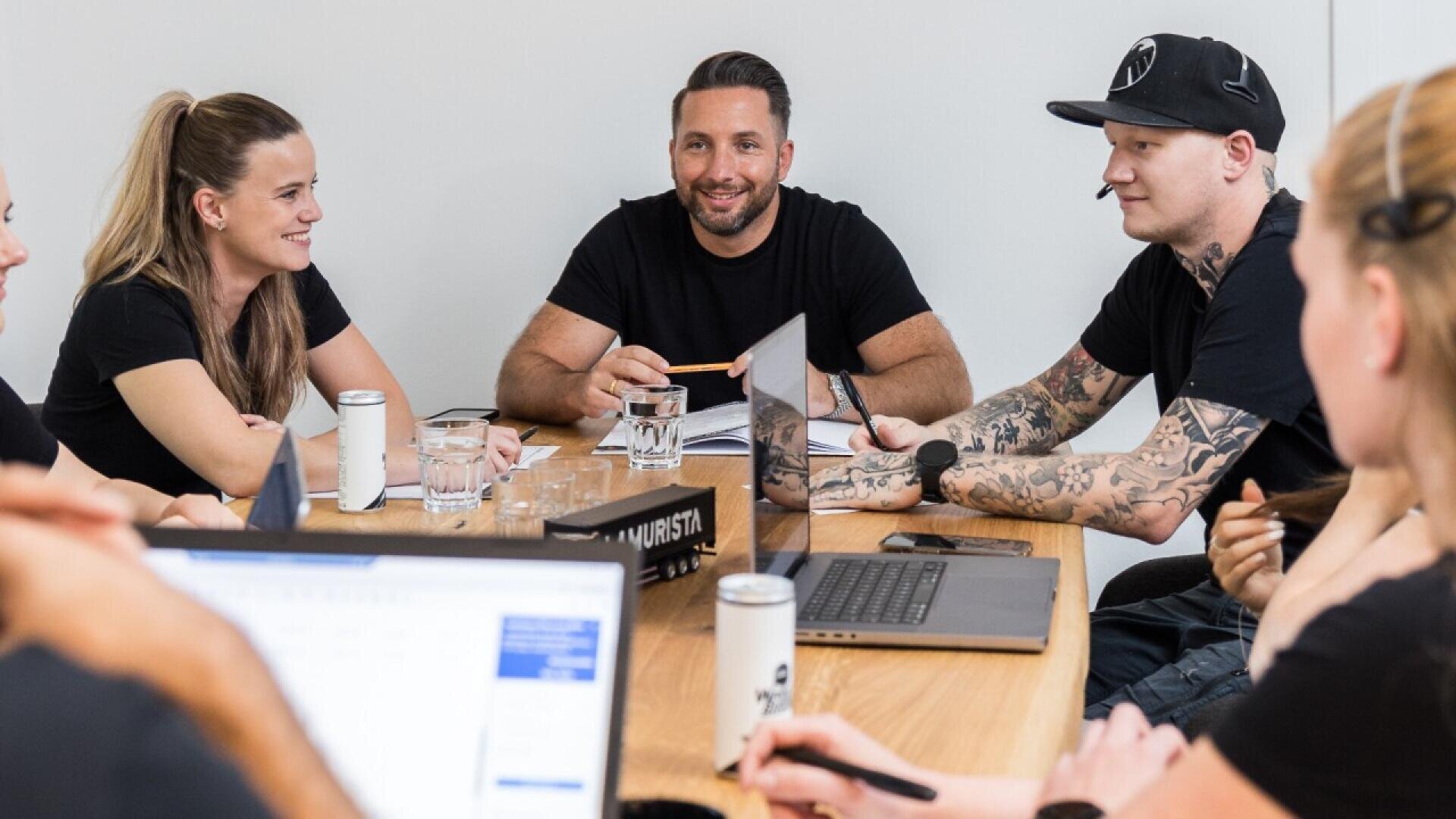 Six people sit around a wooden table in a meeting room, conversing and smiling. Laptops, cans, and glasses of water are on the table, and one man has visible tattoos on his arms.