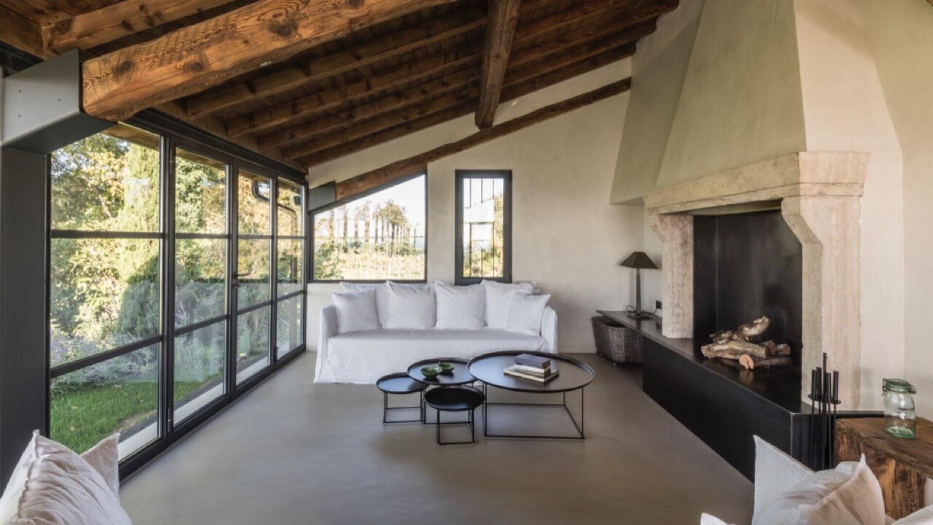 A cozy living room with a rustic wood-beamed ceiling, a white sofa, round black coffee tables, a large stone fireplace, and floor-to-ceiling windows overlooking a green outdoor view.