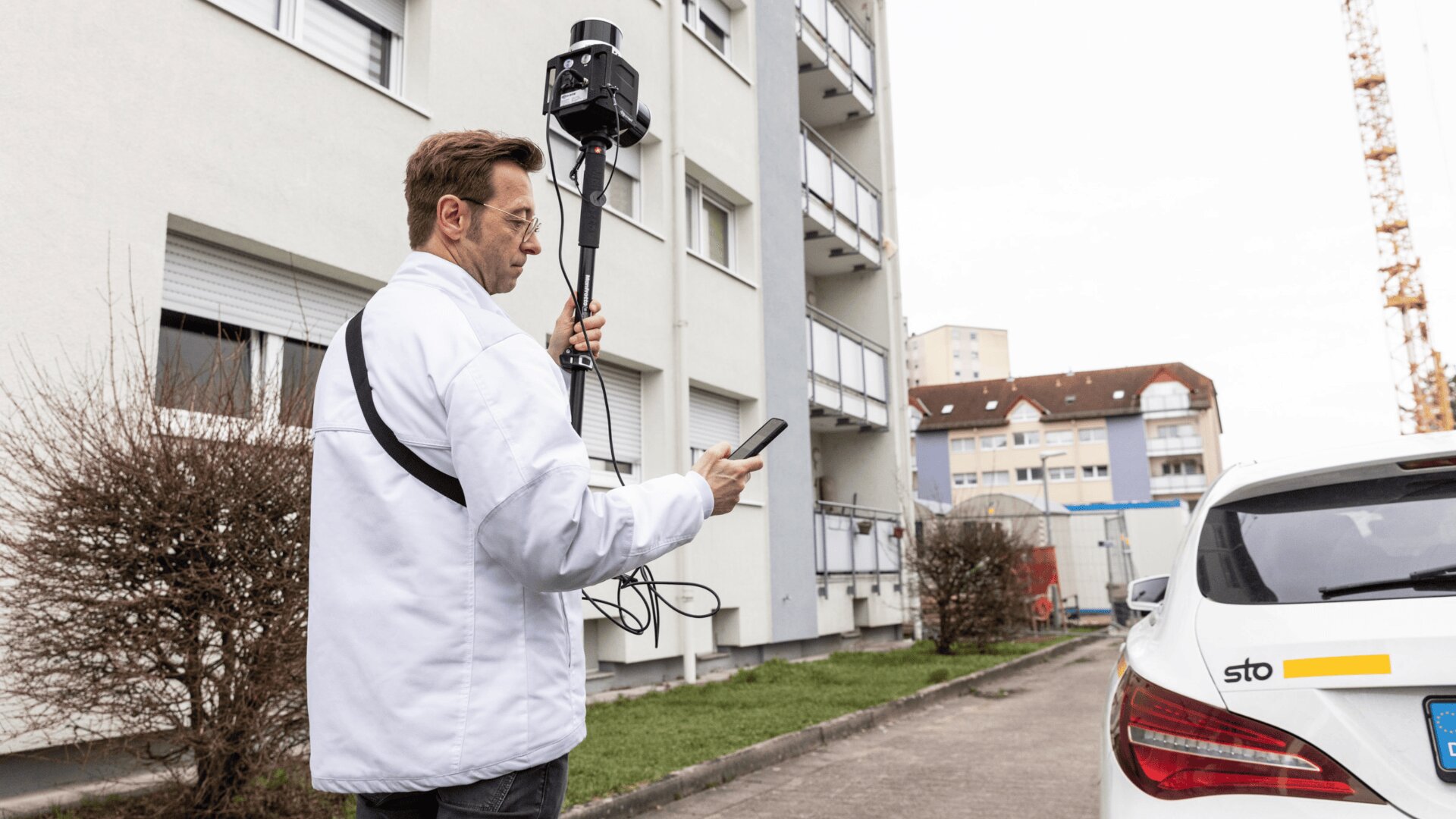 Ein Mann in einer weißen Jacke steht neben einem weißen Auto und einem Wohnhaus, hält ein großes Kameragerät in der Hand und schaut auf sein Smartphone. Es scheint, als würde er im Freien eine Untersuchung oder Inspektion durchführen.
