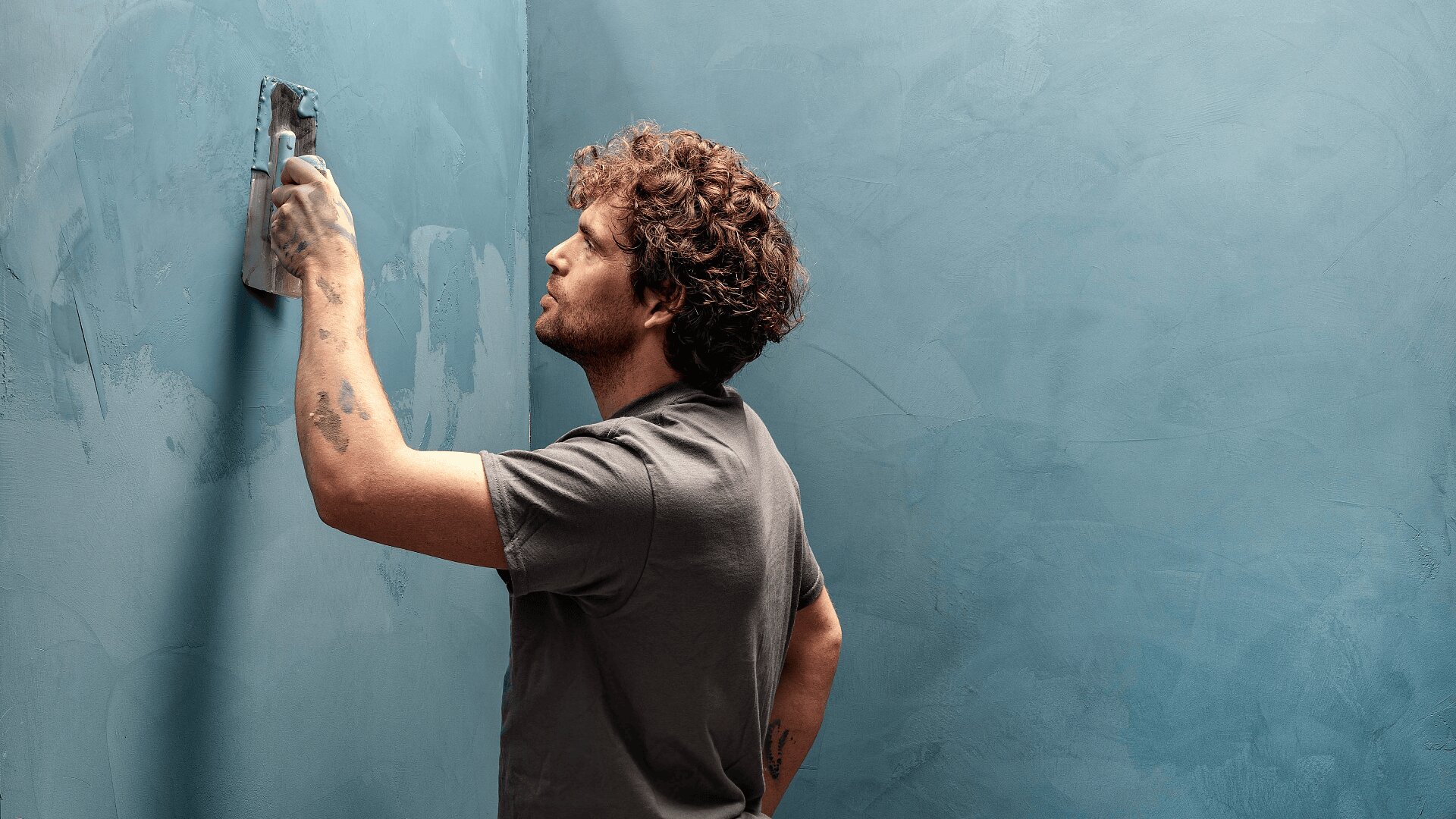 Ein Mann mit lockigem Haar verteilt mit einer Kelle Gips oder Farbe auf einer strukturierten blauen Wand und konzentriert sich auf seine Arbeit. Seine Kleidung und Hände weisen einige Farbflecken auf.