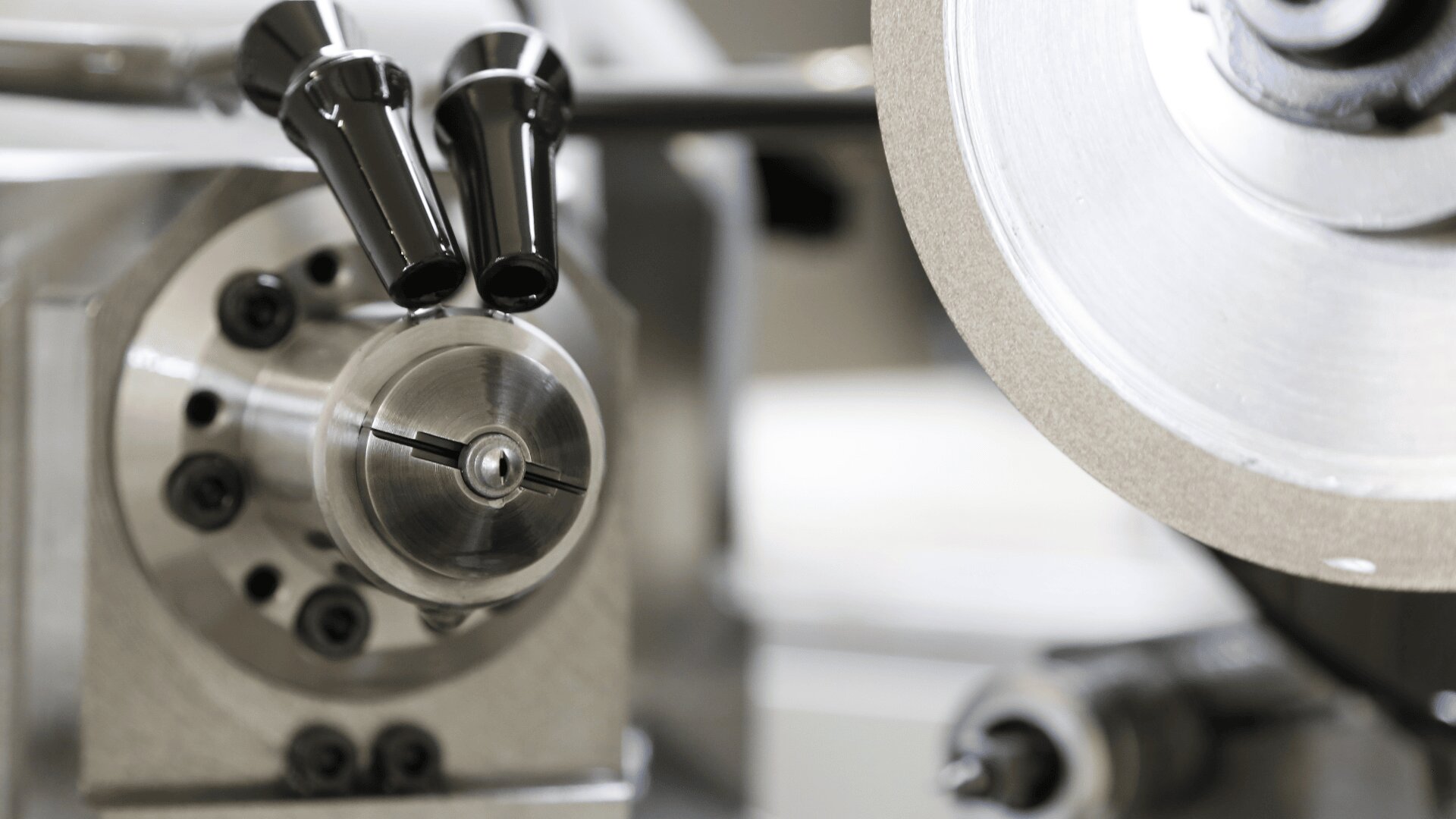 Nahaufnahme einer Präzisionsdrehbank für Metall mit einem runden Spannfutter, zwei schwarzen Hebeln und einer großen rotierenden Schleifscheibe, die alle zum Bearbeiten und Formen von Metallteilen verwendet werden.
