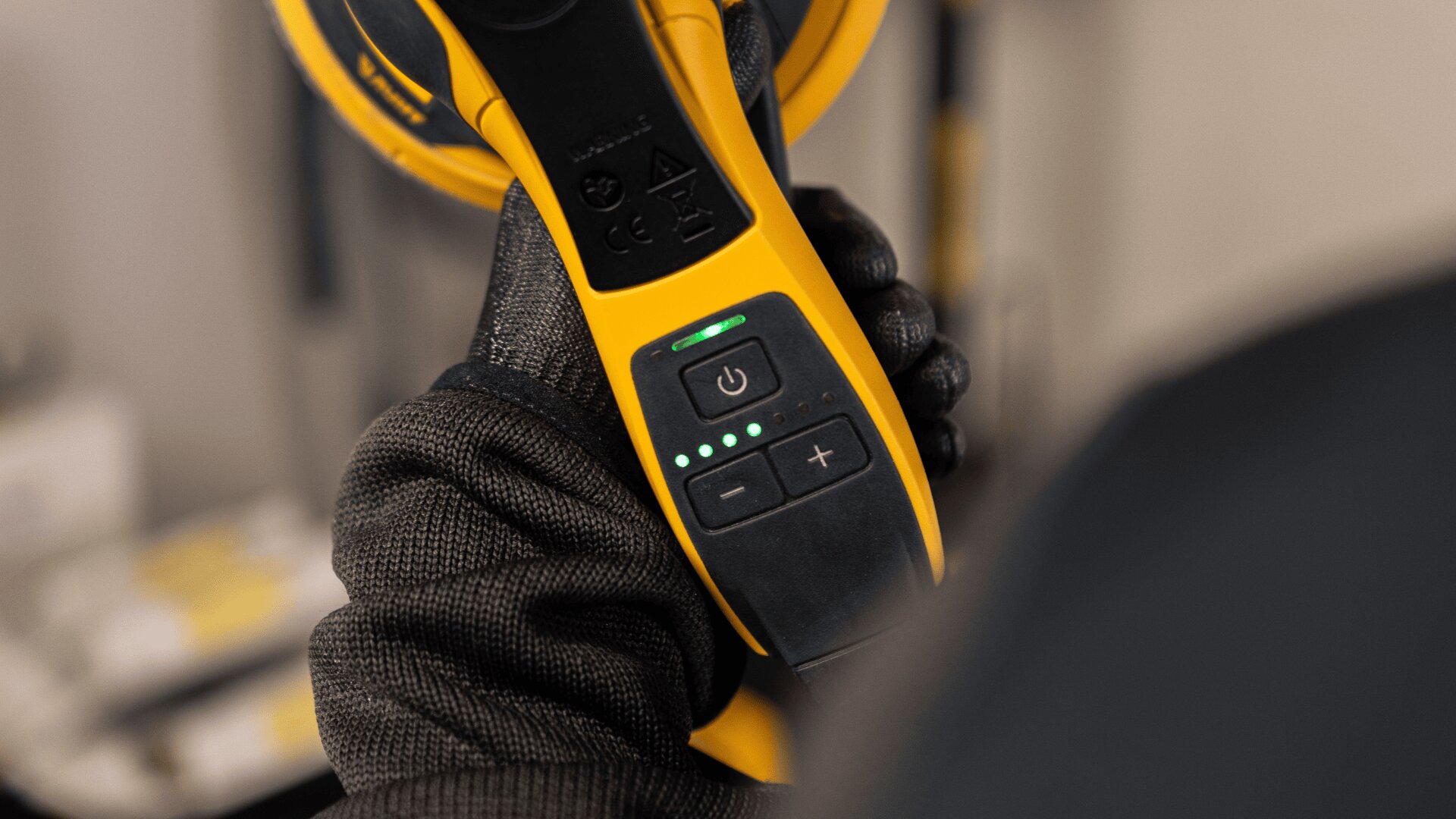 Eine behandschuhte Hand hält ein gelb-schwarzes Elektrowerkzeug und konzentriert sich auf das Bedienfeld mit einem Einschaltknopf, Plus- und Minustasten und vier leuchtenden grünen Kontrollleuchten.