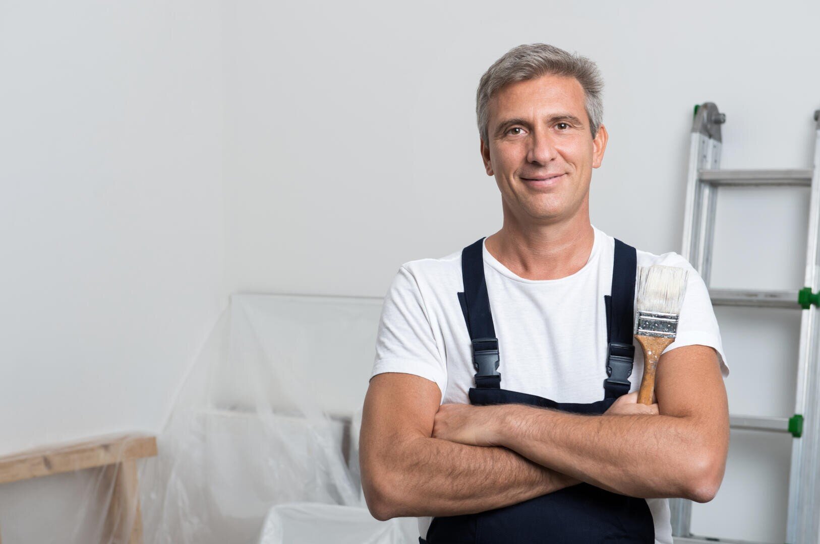 Ein Mann in Overall und weißem T-Shirt steht mit verschränkten Armen da und hält einen Pinsel in der Hand. Er lächelt in einem Raum mit einer Leiter und mit Plastikfolien bedeckten Möbeln, was auf Renovierungs- oder Malerarbeiten hindeutet.