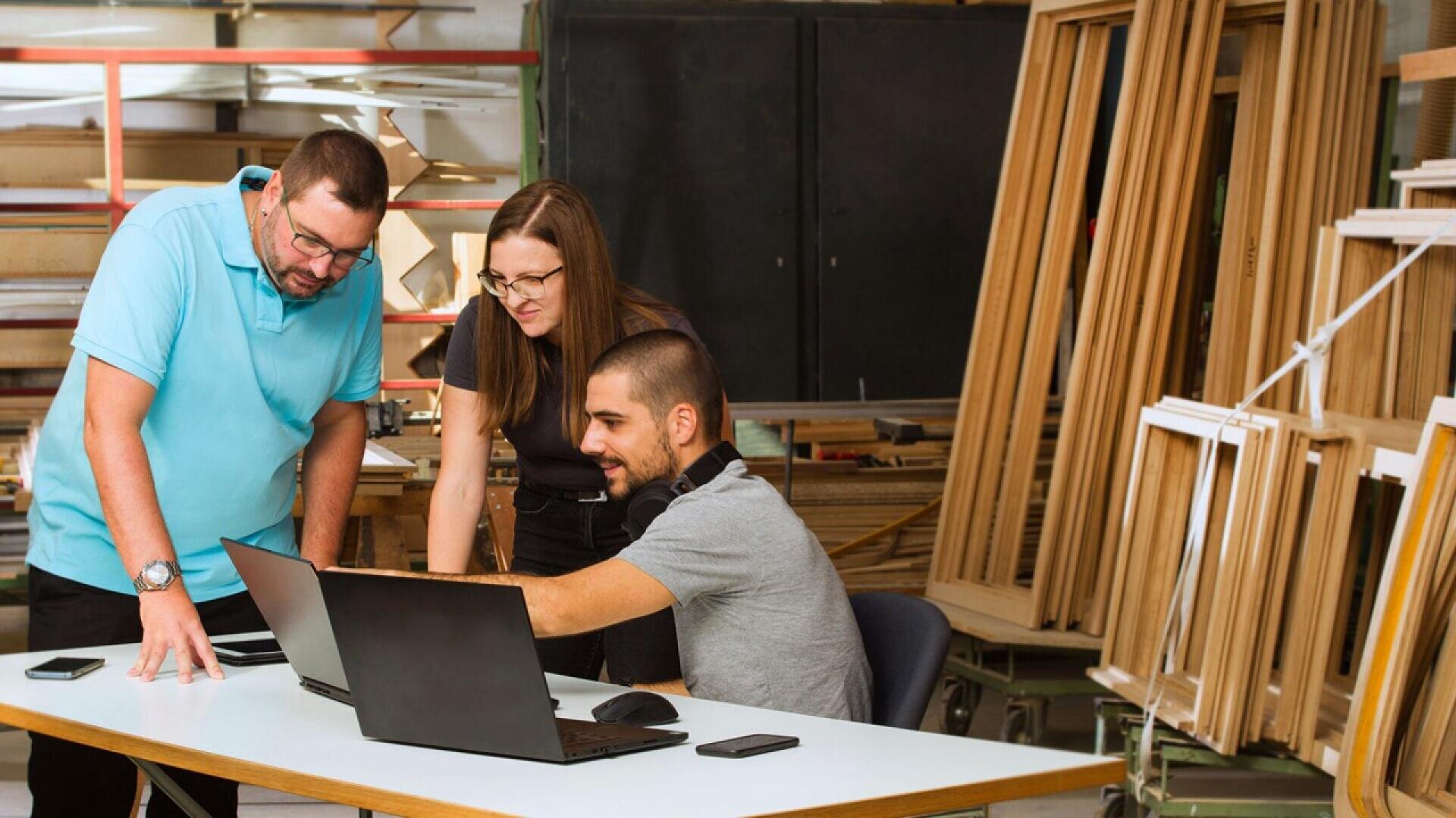 Three people in a workshop look at laptops on a desk; wooden frames and tools are visible in the background, suggesting they are collaborating on a project involving craftsmanship or design.