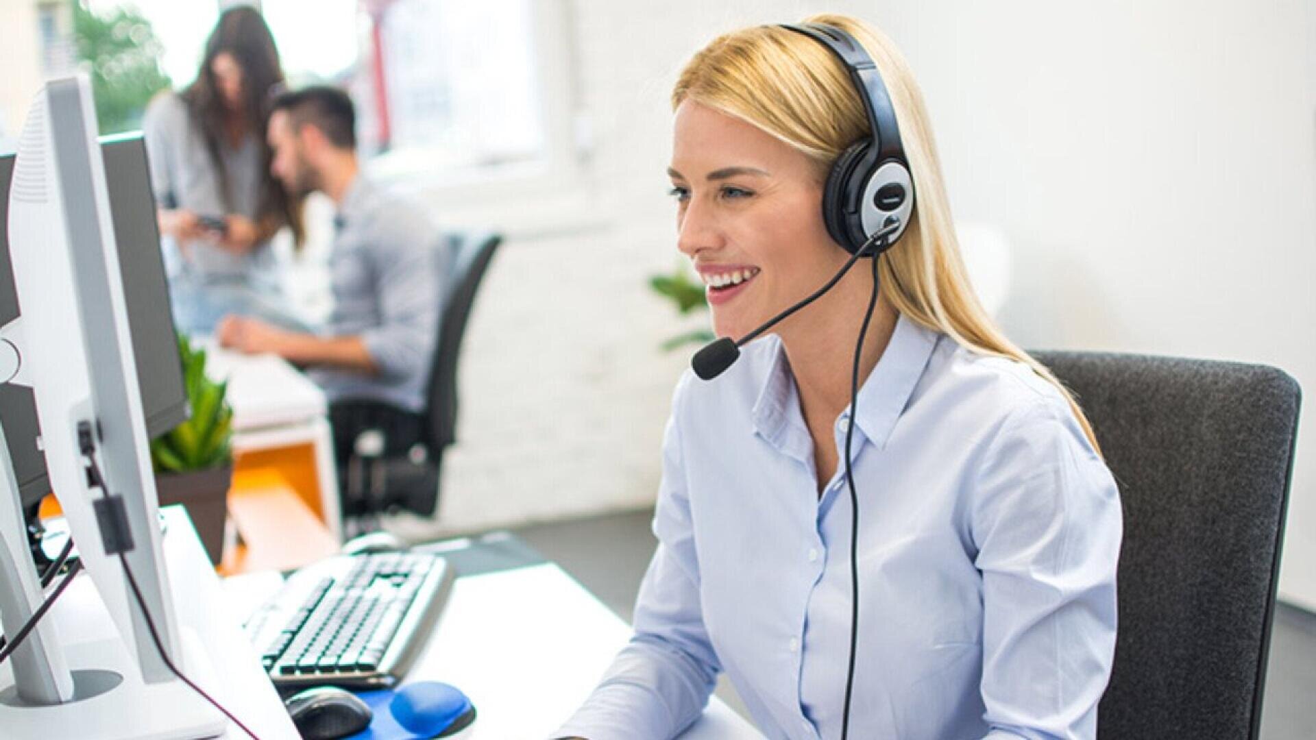 A woman wearing a headset smiles while working at a computer in an office, with two colleagues blurred in the background.