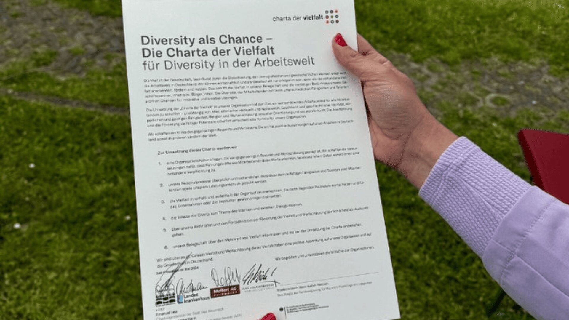 Eine Person hält ein Dokument mit dem Titel Diversity als Chance – Die Charta der Vielfalt für Diversity in der Arbeitswelt und unten mit Unterschriften in der Hand und steht draußen im Gras.