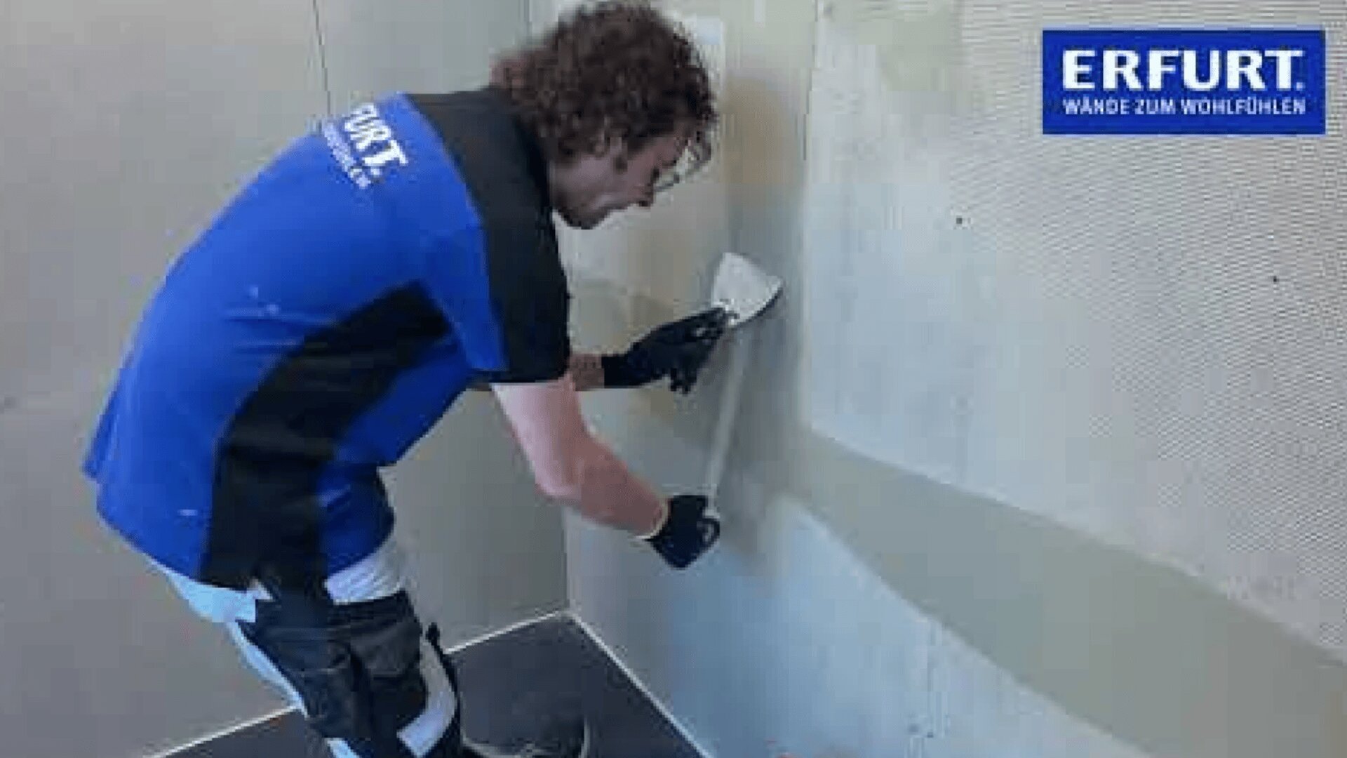 Eine Person mit Arbeitshandschuhen und Erfurt-Shirt trägt mit einem Spachtel Material auf einer Wand auf, in der oberen rechten Ecke ist das Erfurt-Logo zu sehen.