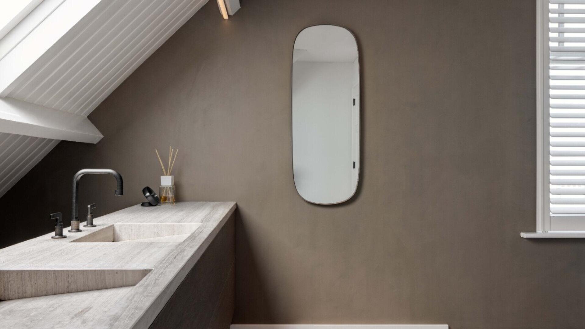 Minimalist bathroom with sloped ceiling, a rectangular stone sink, black faucet, reed diffuser, brown wall, oval mirror, and a window with white blinds letting in natural light.