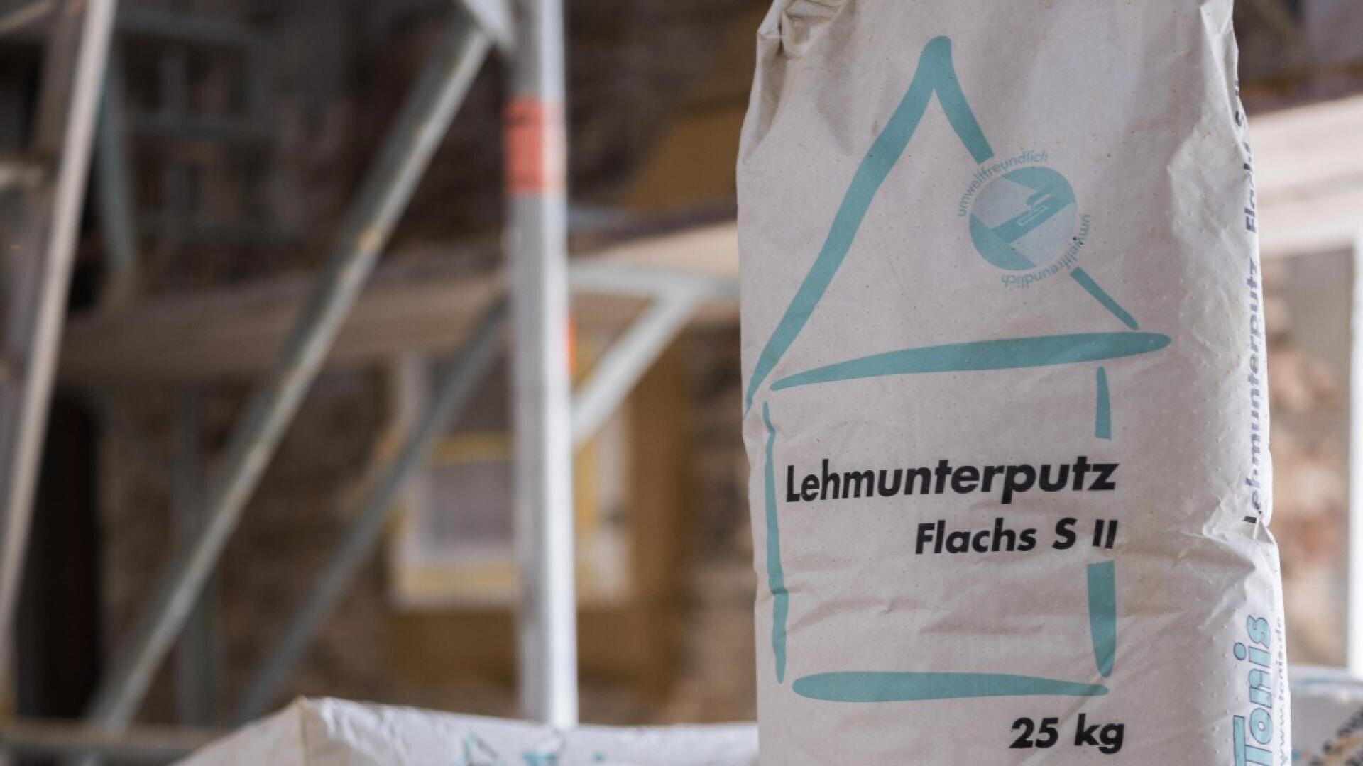 A close-up of a 25 kg bag labeled Lehmunterputz Flachs S II with teal graphics, standing upright at a construction site with scaffolding and blurred background.