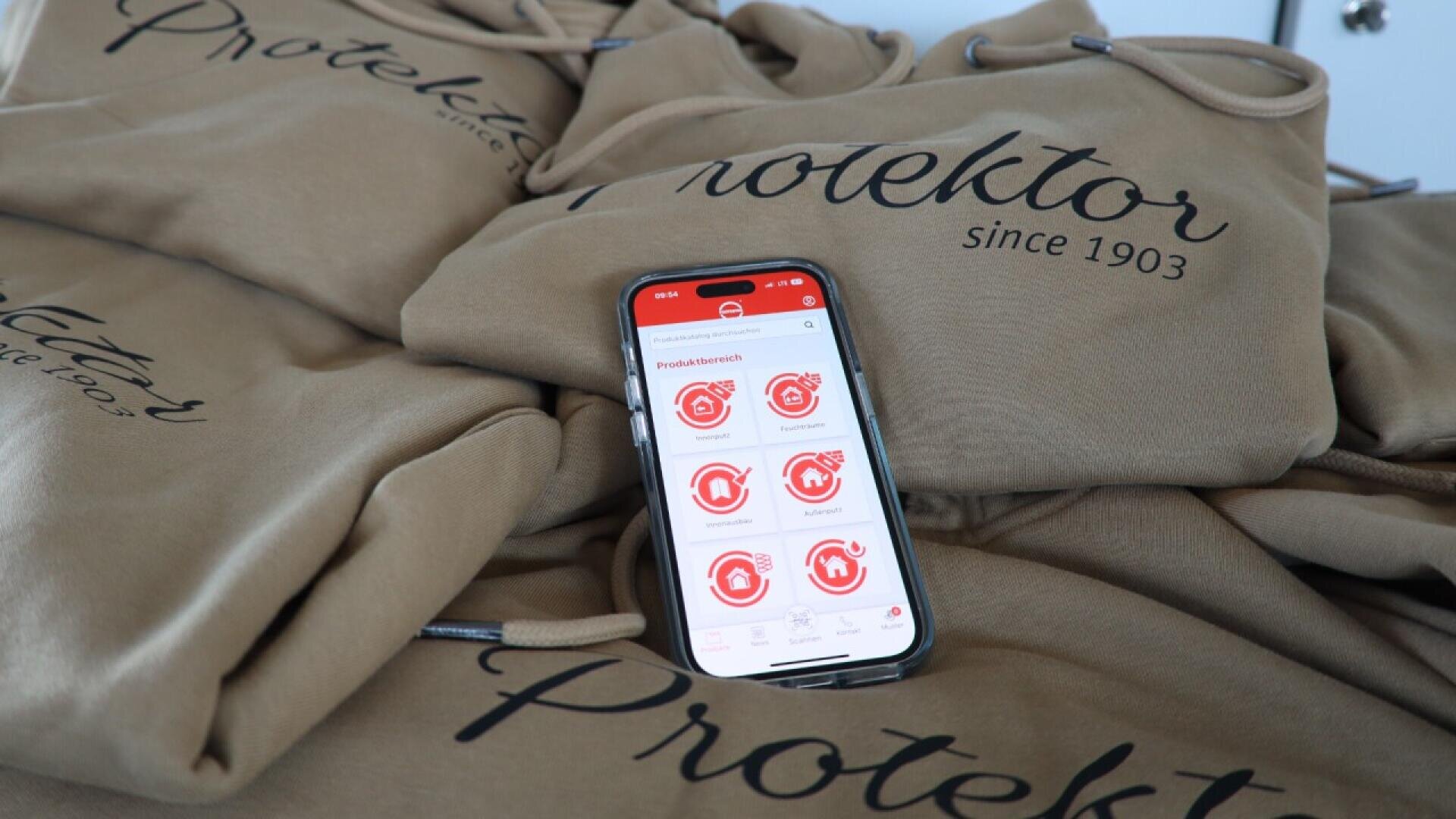 A smartphone displaying an app with red icons rests on a pile of tan hoodies that have Protektor since 1903 printed in black script.