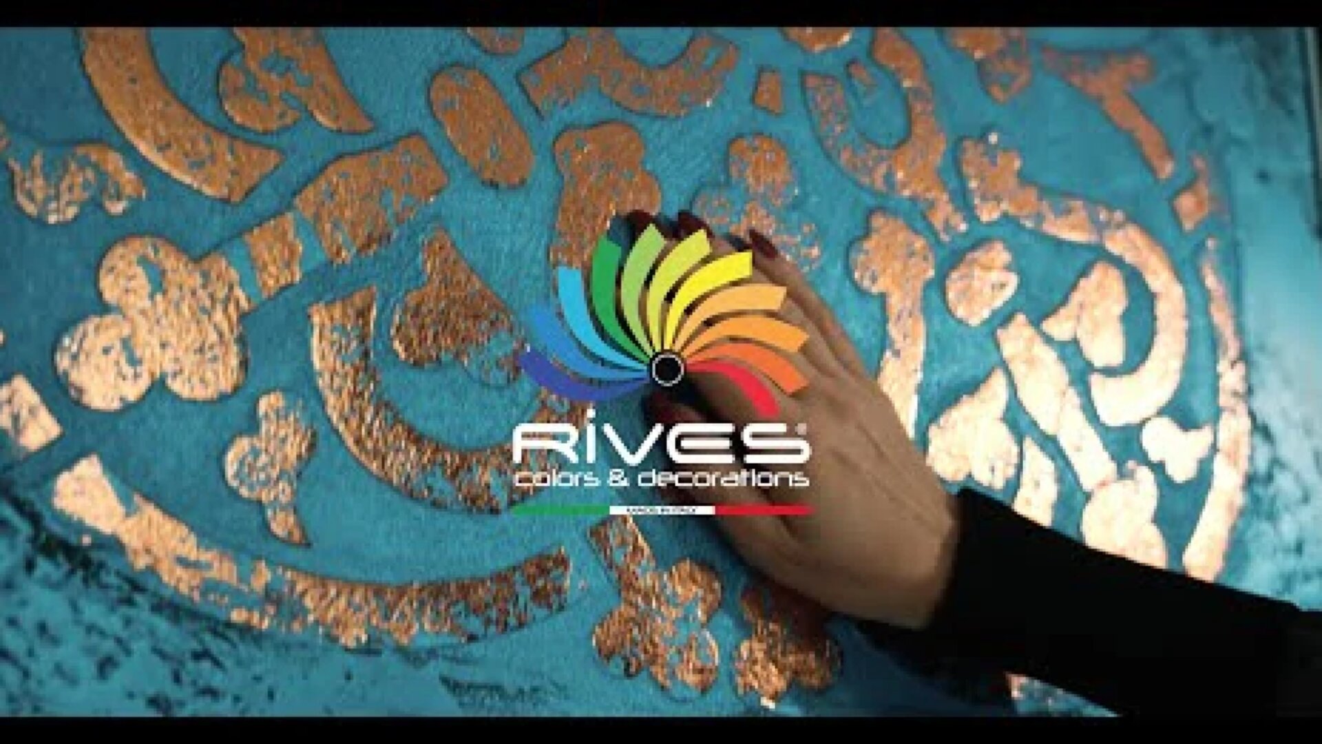 Eine Hand berührt eine blaue Wand mit goldenen Dekormustern. Das Logo von RIVES Colors & Decorations mit einem bunten Wirbel ist zentriert über dem Bild platziert.