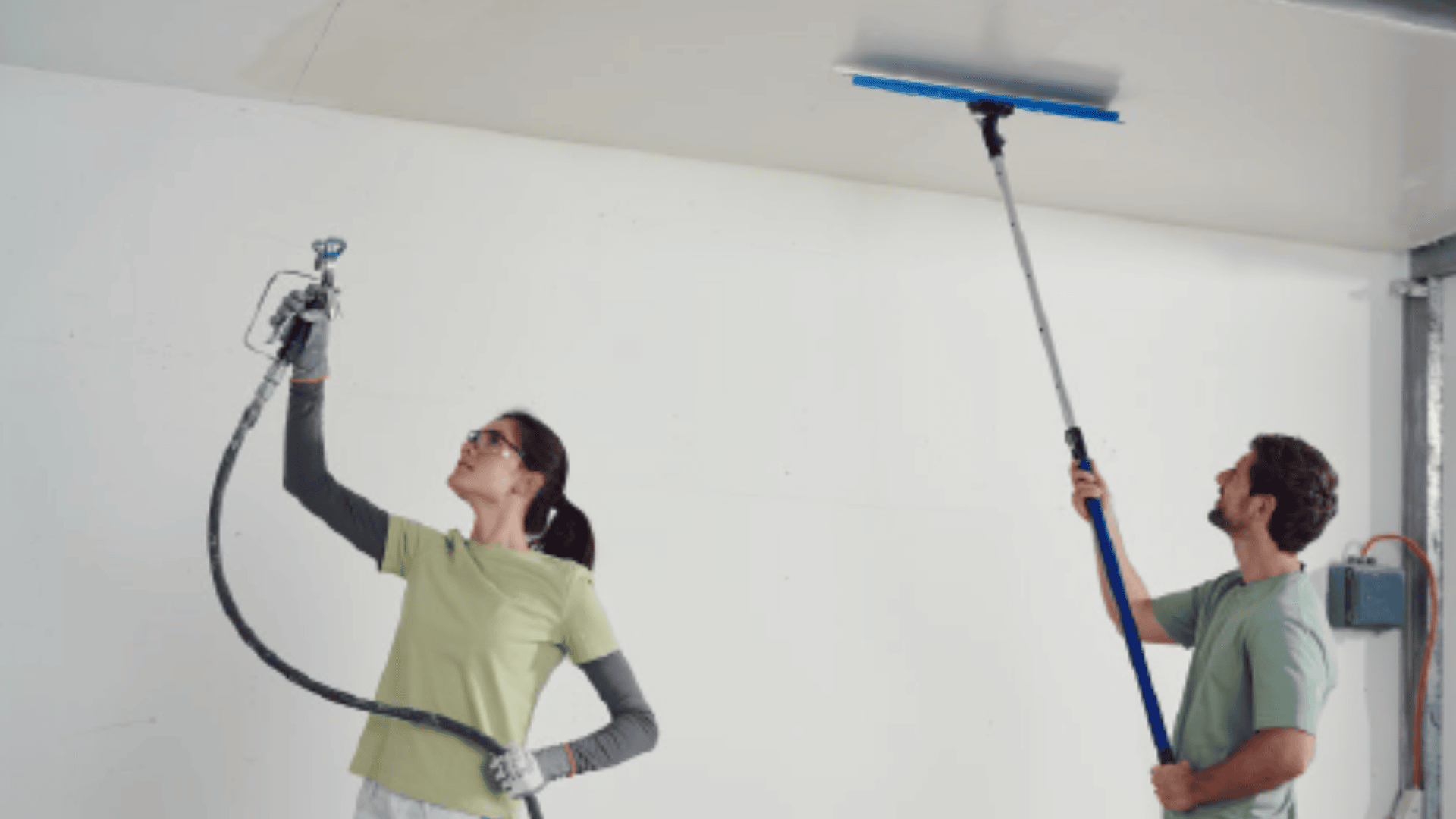 Zwei Personen in Freizeitkleidung arbeiten im Innenbereich; eine spritzt die Decke mit einer Farbpistole ab, während die andere die Decke mit einem langstieligen Werkzeug glättet oder schleift.