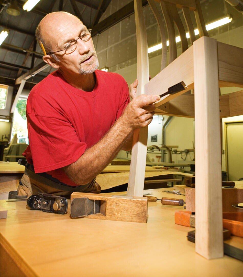 A man in a red shirt carefully uses a chisel to work on a wooden chair in a woodworking shop, surrounded by woodworking tools on a table.