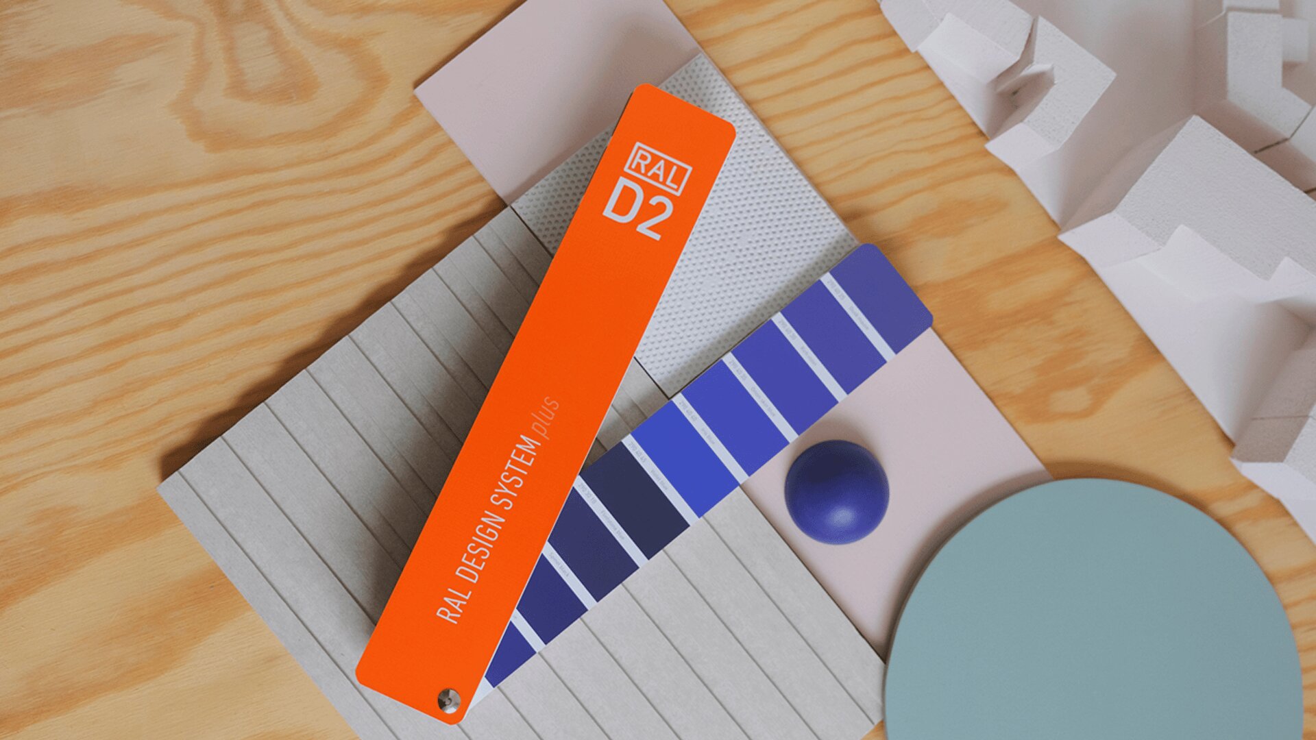Ein orangefarbener RAL D2-Farbfächer liegt diagonal über einem blauen Farbstreifen auf einer Holzoberfläche, um die herum graue, blaue und weiße geometrische Objekte angeordnet sind.
