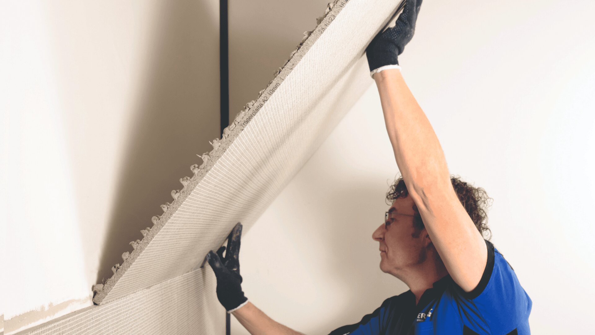 Eine Person mit Handschuhen hebt eine große Gipskartonplatte an und befestigt sie mit Klebstoff an einer Wand.