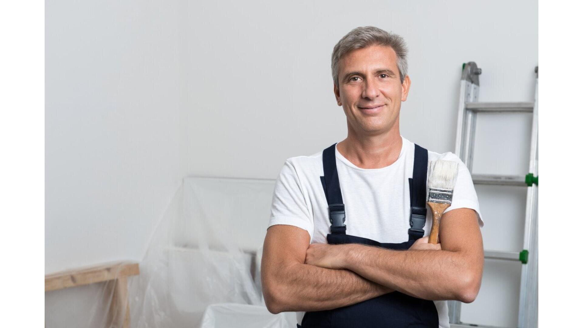 A middle-aged man with gray hair, wearing a white t-shirt and dark overalls, stands with arms crossed and a paintbrush in his pocket. He is smiling, and there is a ladder and covered furniture in the background.