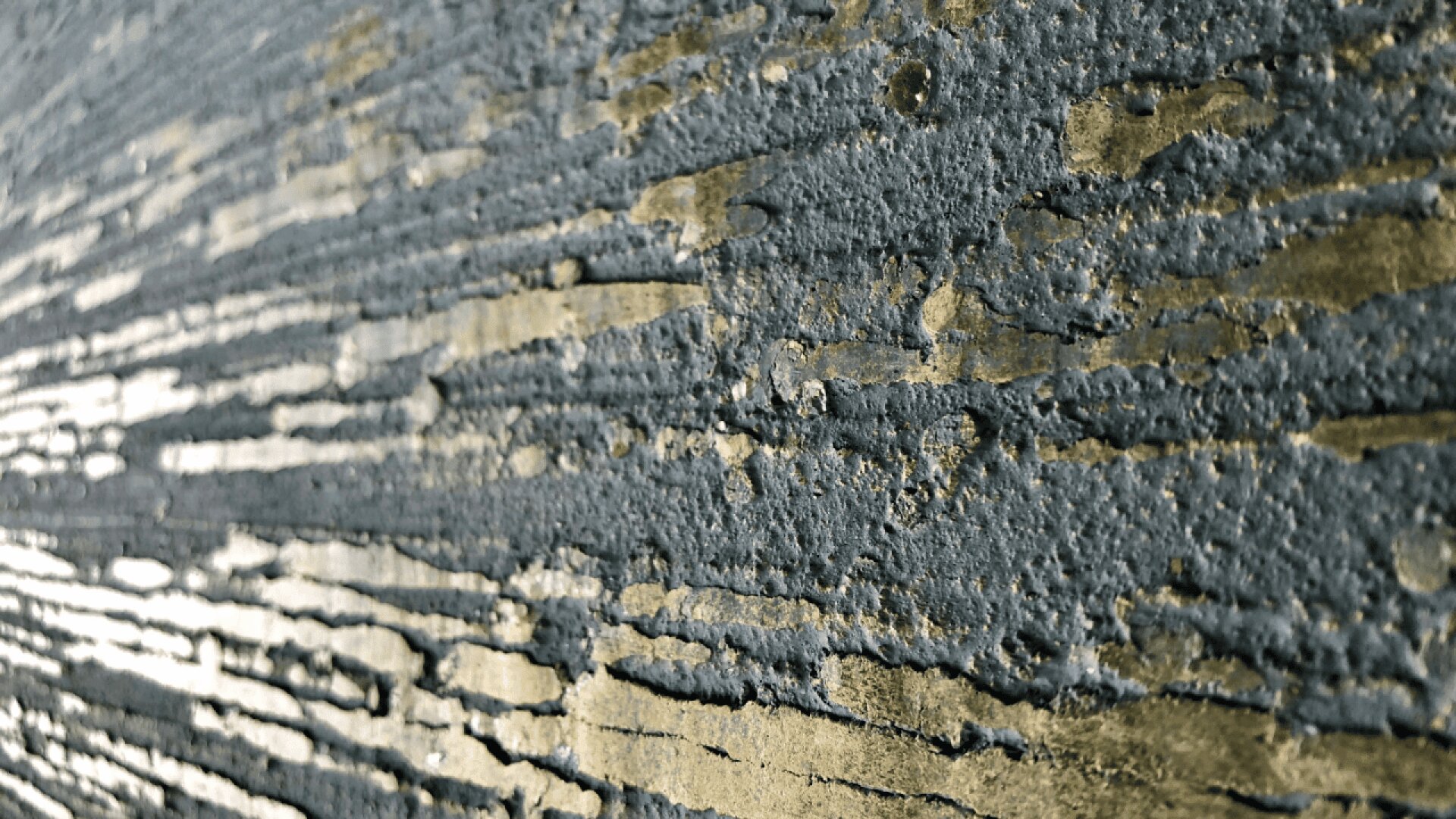 Close-up of a textured wall with horizontal streaks of exposed gold and gray layers, creating an abstract, weathered pattern. Light shines from the left, highlighting the rough, uneven surface.