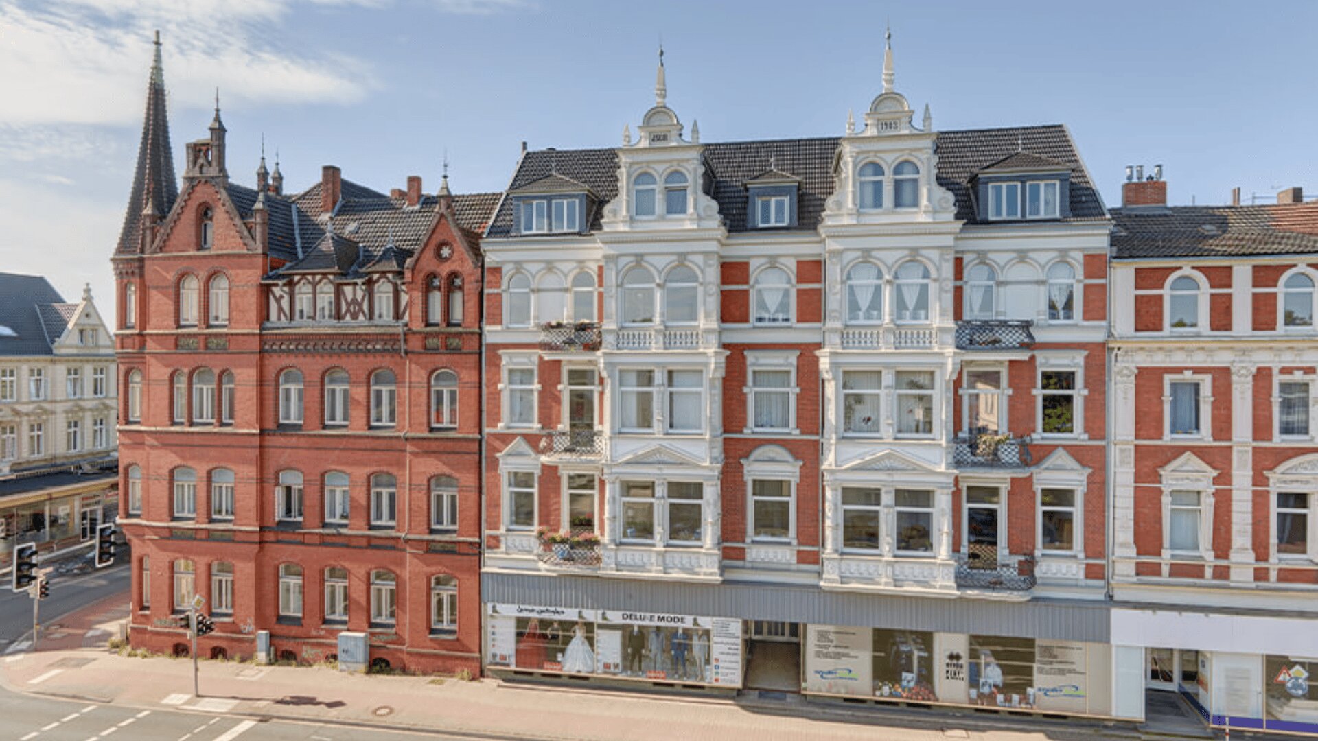 Eine Straßenansicht reich verzierter, historischer europäischer Gebäude mit roten Backstein- und weißen Fassaden, hohen Fenstern, bepflanzten Balkonen und einem spitzen Turm auf der linken Seite unter einem klaren blauen Himmel.