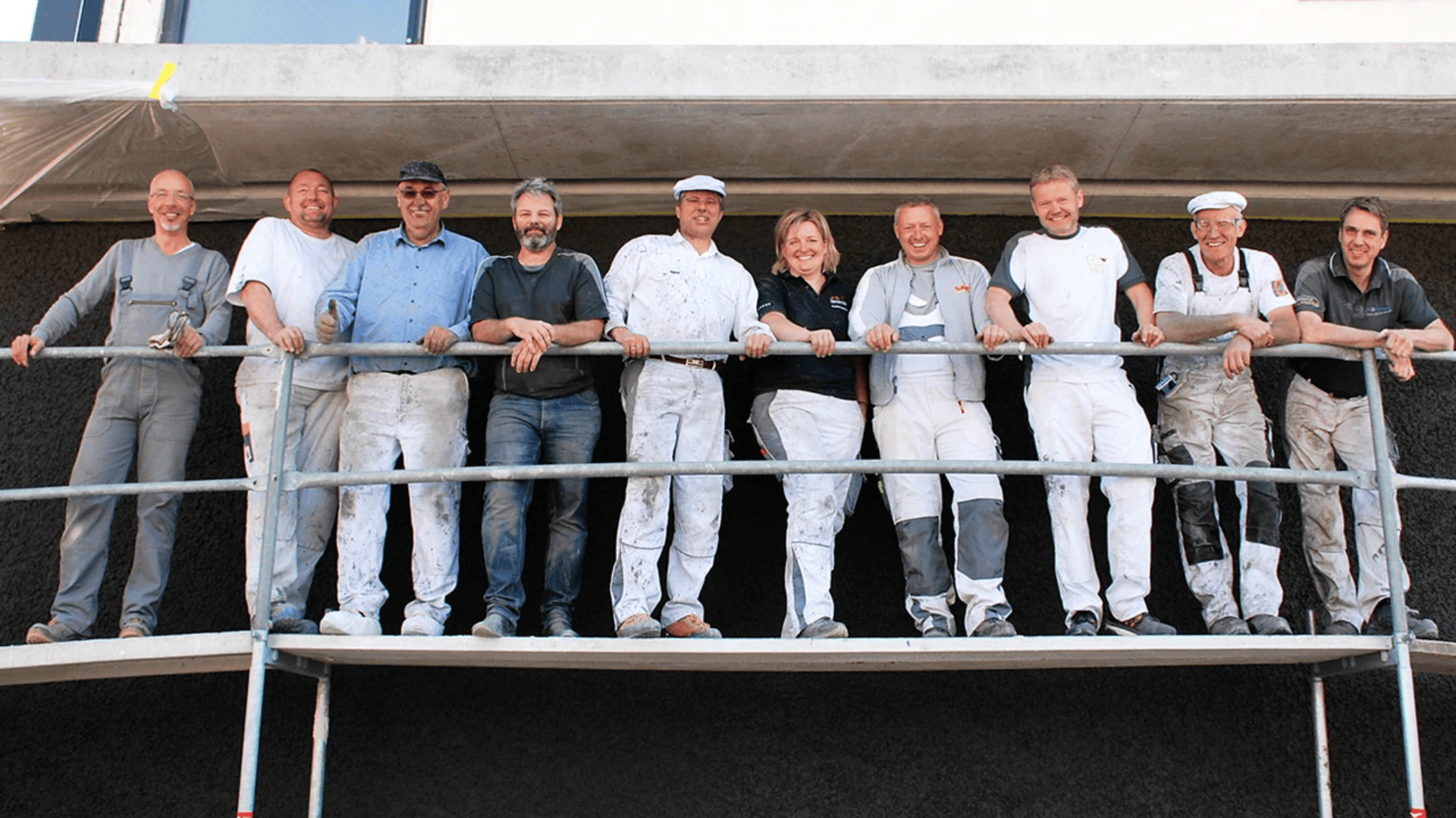 Zehn Menschen, manche in Freizeitkleidung, andere in Arbeitsuniformen, stehen lächelnd zusammen auf einem Gerüst vor einem im Bau befindlichen Gebäude.