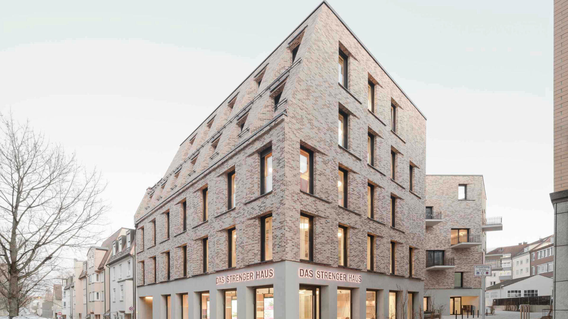 Ein modernes mehrstöckiges Gebäude mit schrägem Dach und großen Fenstern, erbaut aus hellen Ziegeln. Im Erdgeschoss steht das Schild „DAS STRECKER HAUS“. Das Gebäude steht an einer Straßenecke.