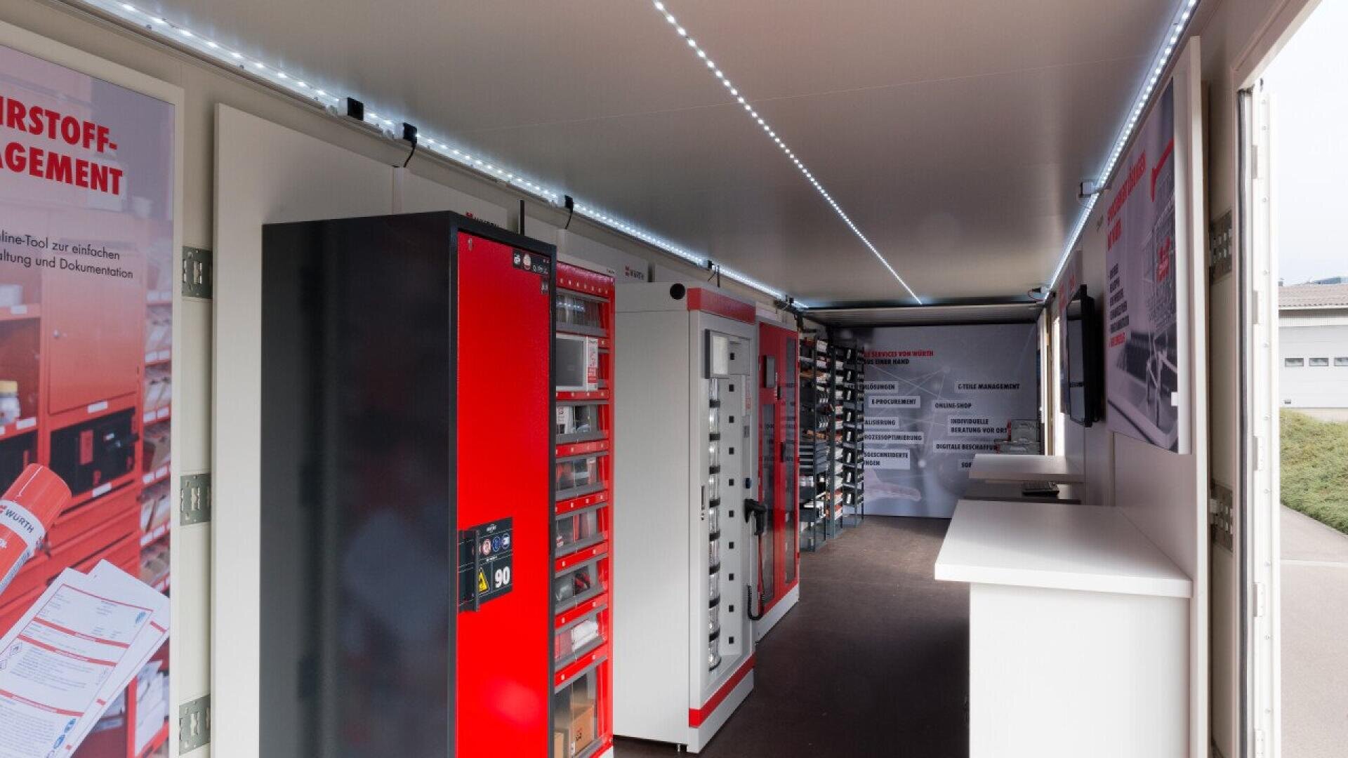 Interior of a mobile unit featuring red and black vending machines, white countertops, wall displays, and a ceiling lined with LED strip lights, used for tool or material management and distribution.