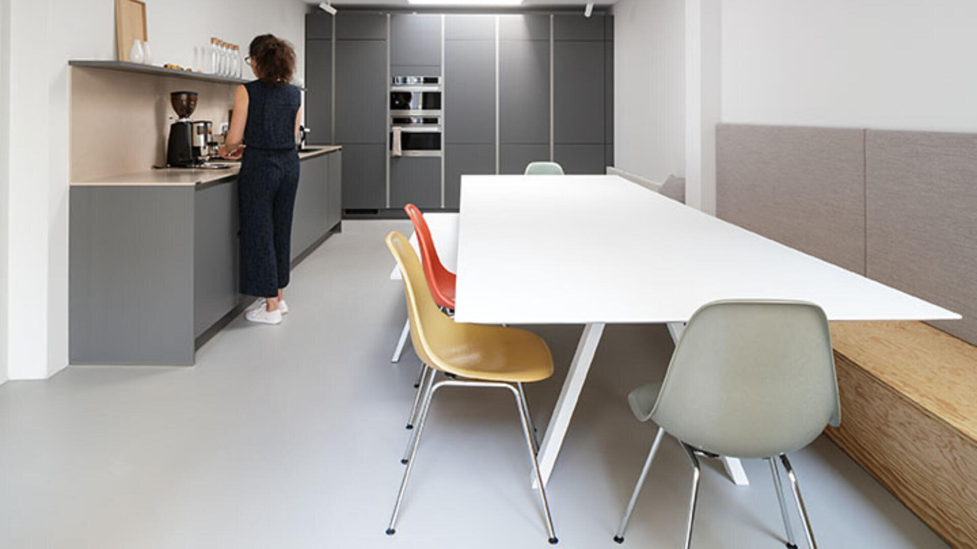 Eine Frau steht an einer Küchentheke in einer modernen, minimalistischen Küche mit grauen Schränken. Im Vordergrund ist ein weißer Esstisch von bunten Stühlen und einer eingebauten Holzbank umgeben.