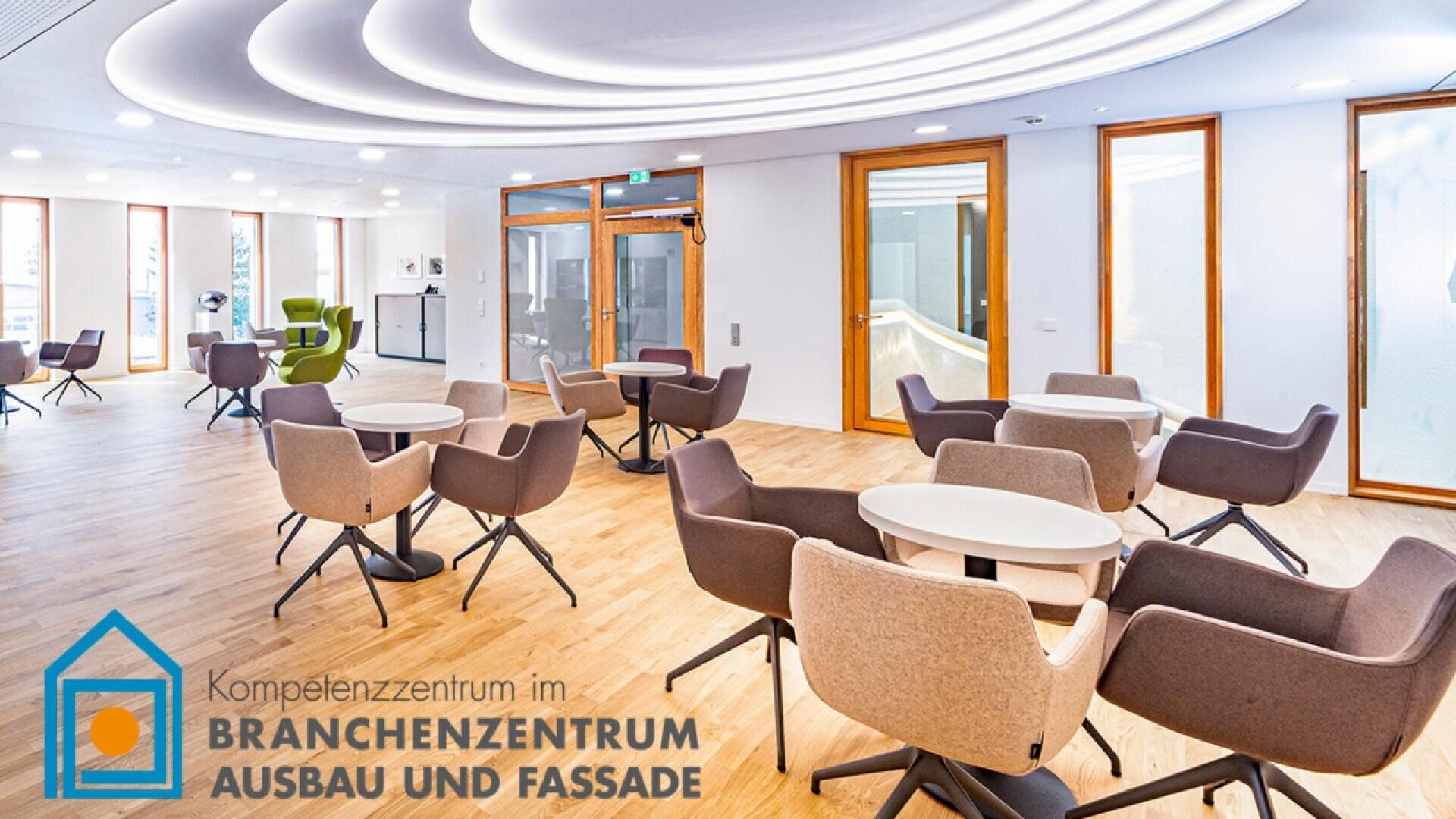 Modern, spacious lounge area with round tables and gray chairs on light wood flooring. Large windows and glass doors let in natural light. Logo in German at the bottom left reads BRANCHENZENTRUM AUSBAU UND FASSADE.