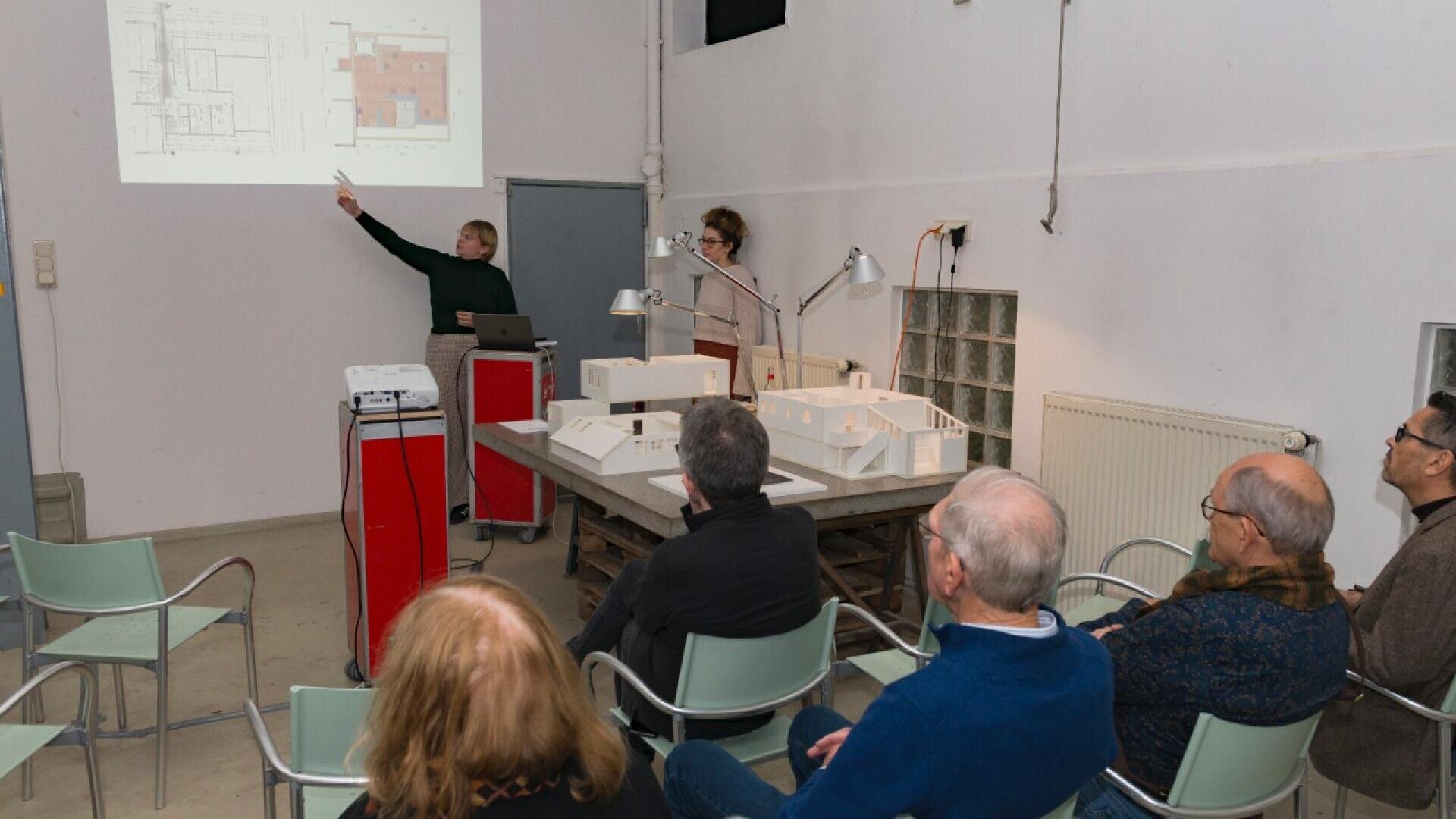 A person points at architectural plans projected on a screen while another stands nearby; several people seated in chairs watch, with scale models of buildings on a table in front.
