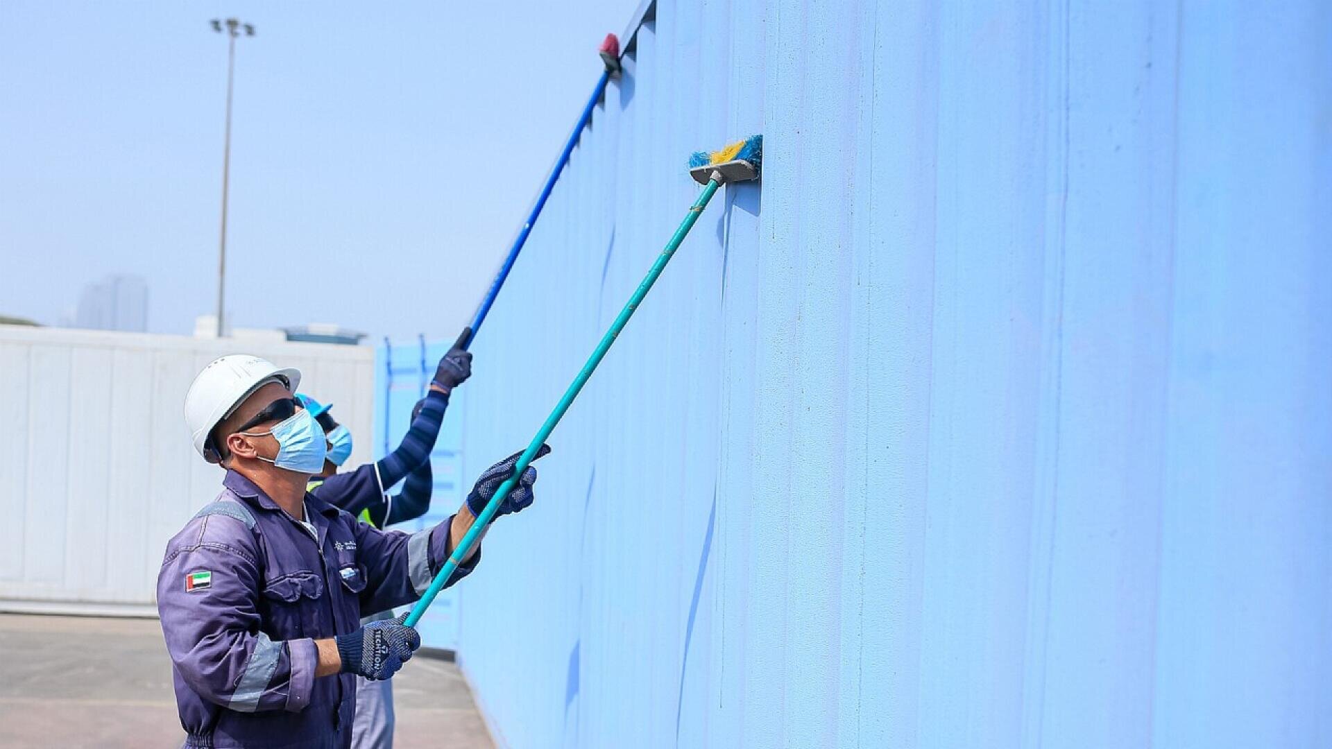 Two workers wearing helmets, masks, and uniforms use long-handled rollers to paint a large exterior wall blue on a sunny day.