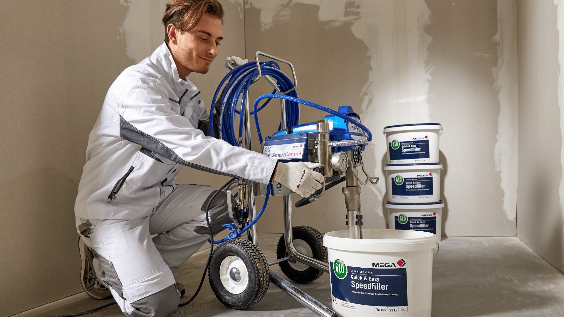 Eine Person in weißer Arbeitskleidung bedient eine Farbspritzmaschine und bereitet sich in einem Raum im Bau darauf vor, Farbe aus einem großen weißen Eimer mit der Aufschrift „Speedfiller“ zu verwenden. Weitere Farbeimer sind in der Nähe gestapelt.