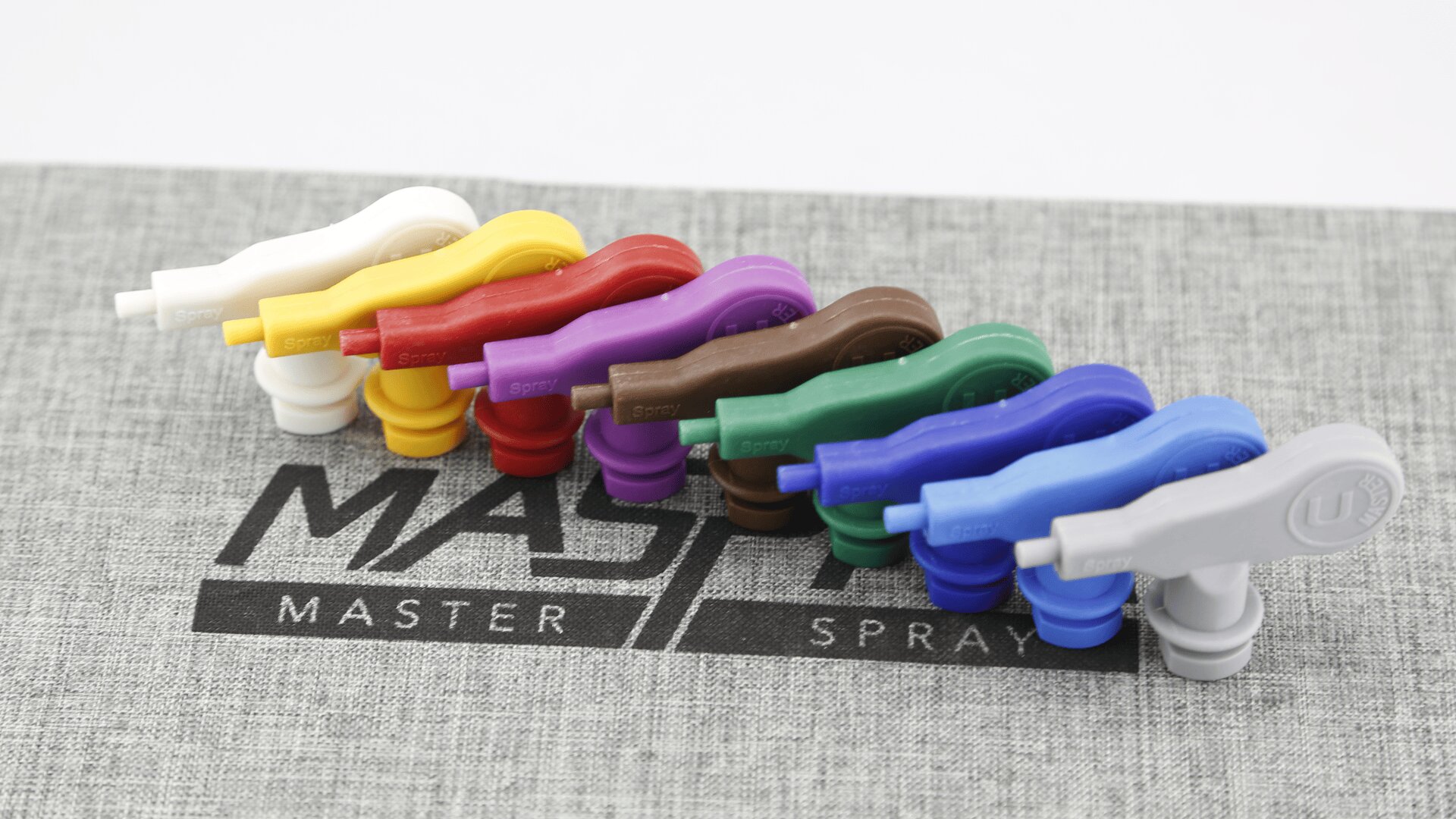 Acht bunte Sprühdüsen aus Kunststoff sind in einer Reihe auf einer grauen, strukturierten Oberfläche angeordnet, darunter ist teilweise der Schriftzug „MASTER SPRAY“ zu sehen.