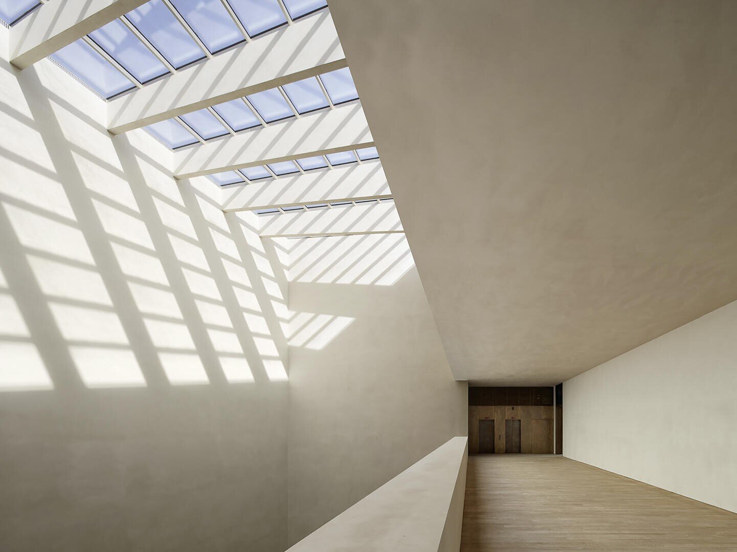 Ein geräumiger, minimalistischer Innenraum mit beigen Wänden und Böden. Ein großes, schräges Oberlicht wirft geometrische Schatten und Sonnenlicht auf die Oberflächen. Am anderen Ende des Raumes sind Holztüren sichtbar.