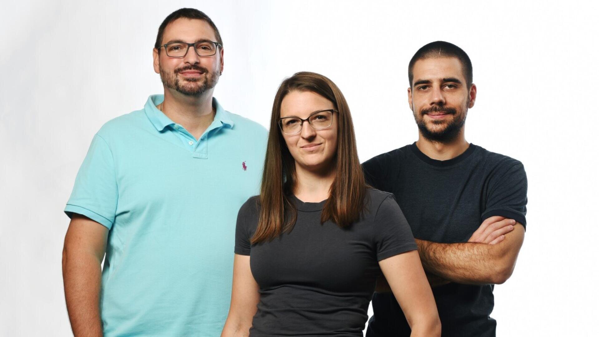 Three people stand together against a white background. The person on the left wears glasses and a light blue polo shirt, the person in the middle has glasses and a gray shirt, and the person on the right has a dark t-shirt and crossed arms.