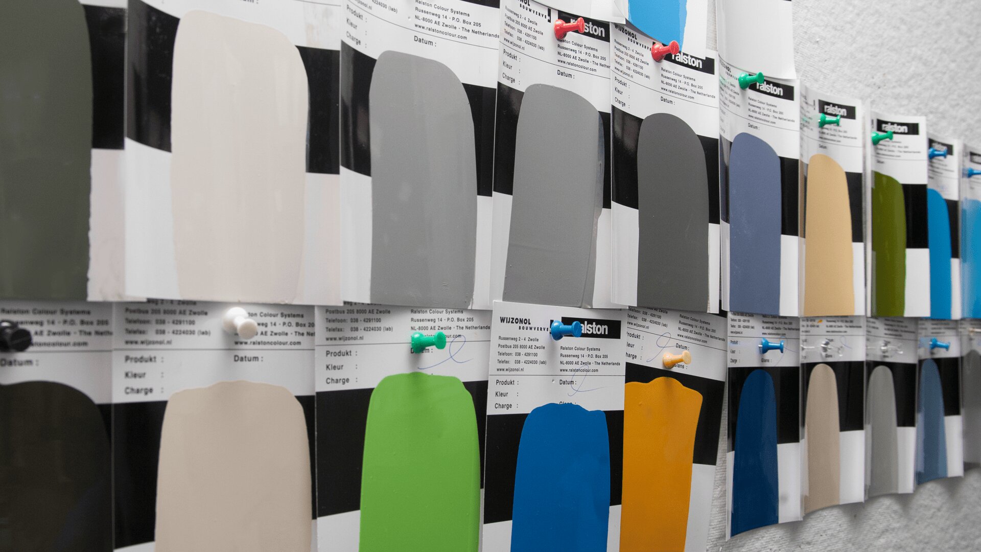 An einer Wand hängen Reihen von Farbmustern, die jeweils mit Reißzwecken befestigt sind. Die Muster zeigen verschiedene Farben, darunter Grau, Blau, Beige, Gelb, Grün und Schwarz.