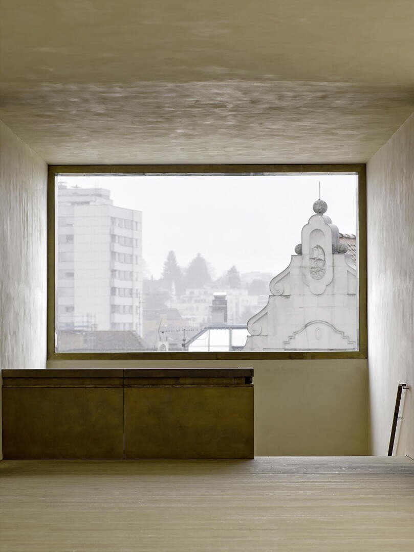Minimalistisches Interieur mit beigen Wänden und einem großen rechteckigen Fenster. Es bietet einen Blick auf die Stadt mit modernen Gebäuden und einer kunstvollen historischen Fassade. Unter dem Fenster steht ein schlichter Schrank.