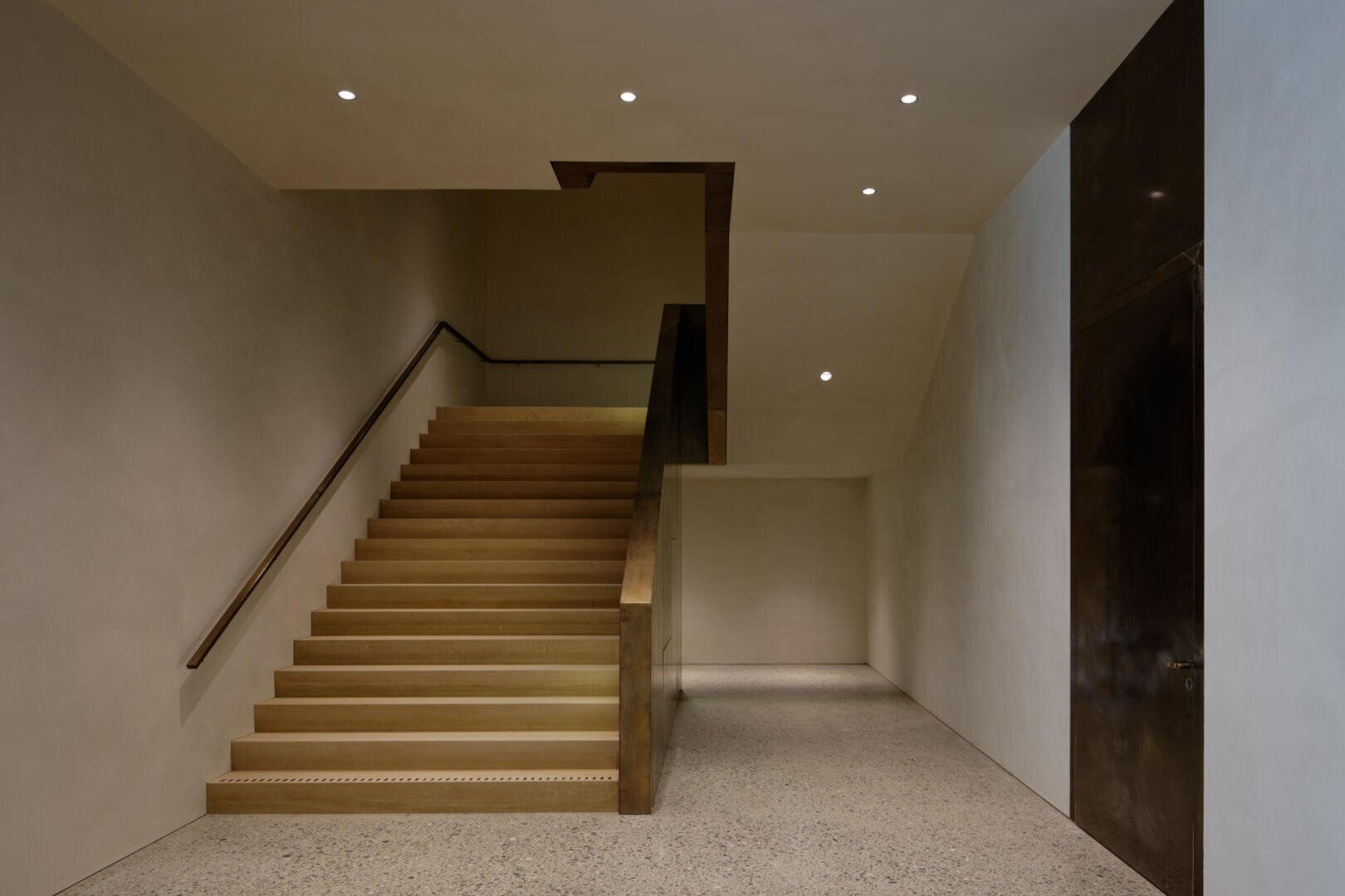 Ein minimalistisches Interieur mit einer breiten Holztreppe mit Metallhandlauf, beigen Wänden, einem polierten Terrazzoboden und eingelassenen Deckenleuchten schafft eine klare, moderne Atmosphäre.