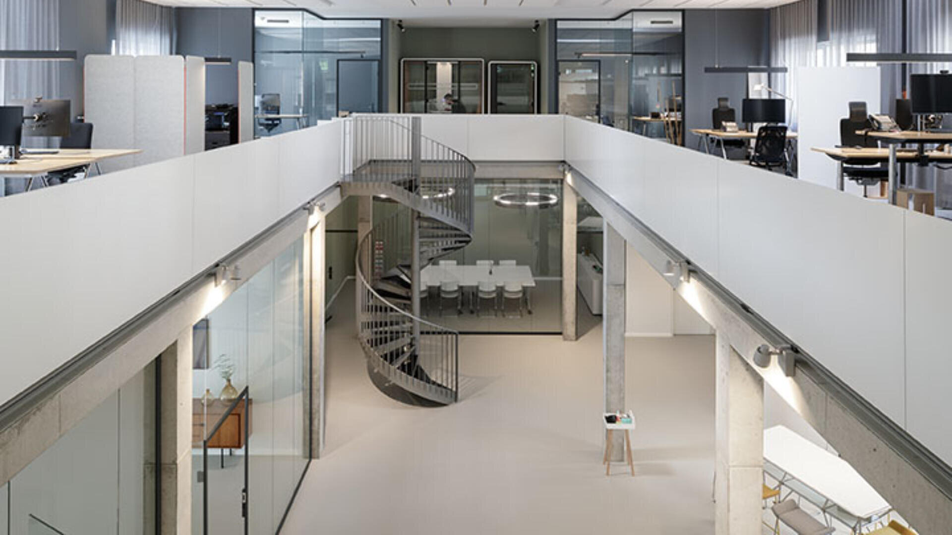 Modernes Bürointerieur mit offenem Mittelbereich, verglasten Besprechungsräumen, einer Wendeltreppe und Arbeitsplätzen im oberen Zwischengeschoss. Der Raum ist hell und mit minimalistischen, modernen Designelementen gestaltet.