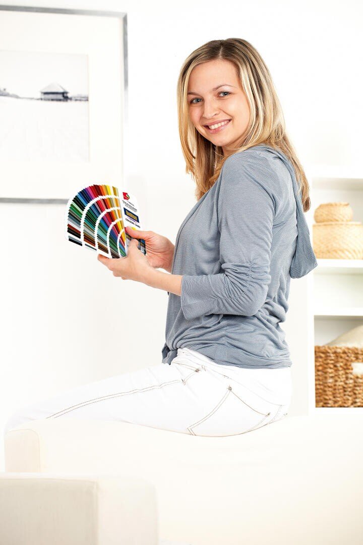 A smiling woman with blonde hair sits sideways on a white couch, holding a color swatch fan. She wears a light gray top and white pants; the background is bright with minimalistic decor and framed art.