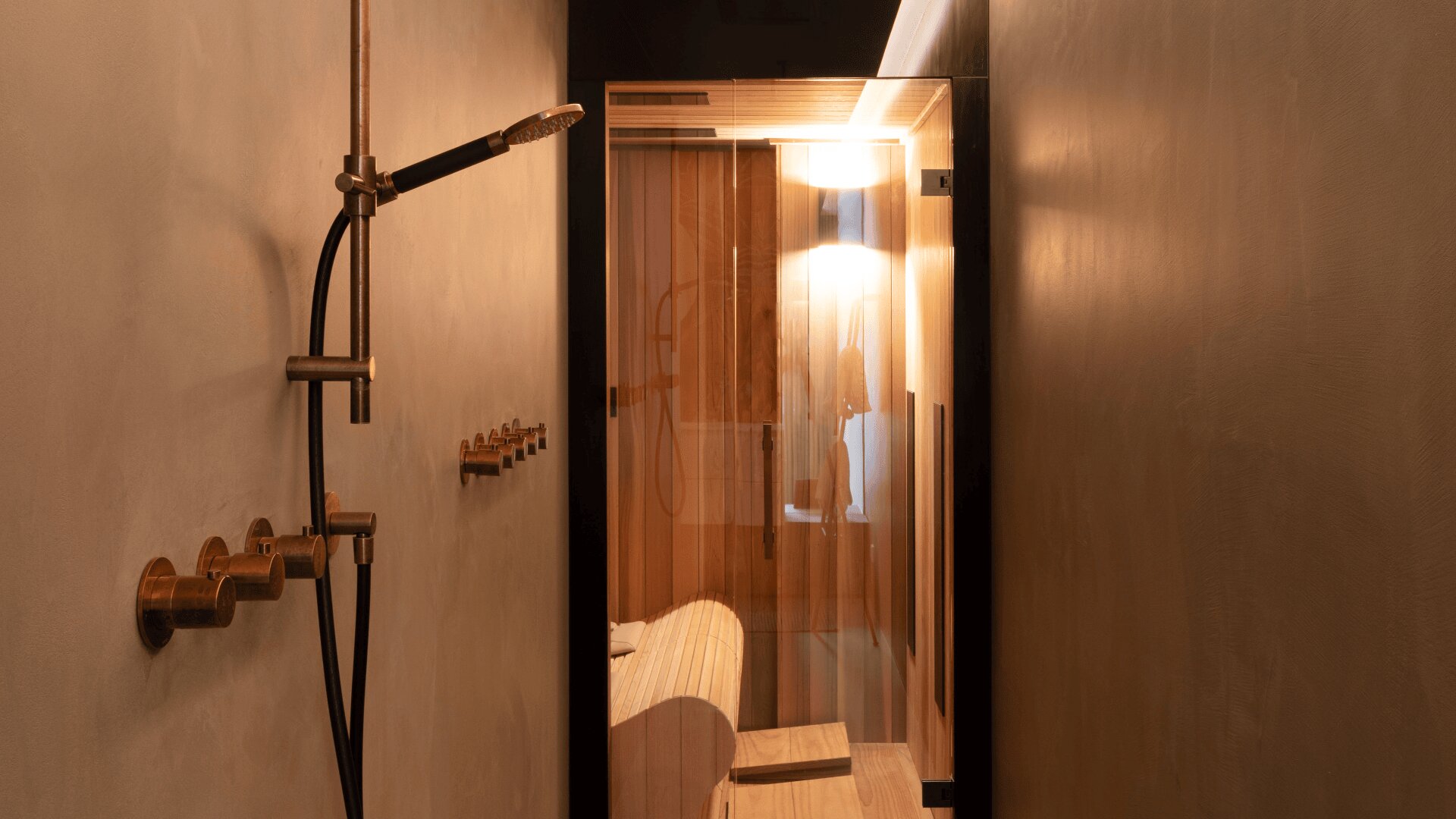 Ein modernes, minimalistisches Badezimmer mit einer bronzenen Duschvorrichtung auf der linken Seite und einem verglasten Saunaraum mit warm beleuchteten Holzbänken am Ende eines schmalen Flurs.