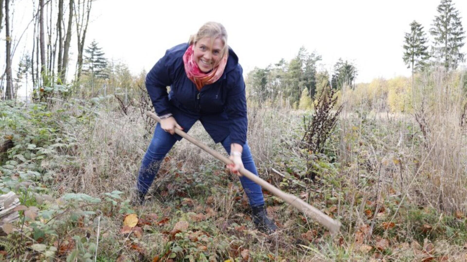 A smiling woman in a scarf and jacket uses a hoe to dig in an overgrown, autumnal garden surrounded by dried plants and trees.