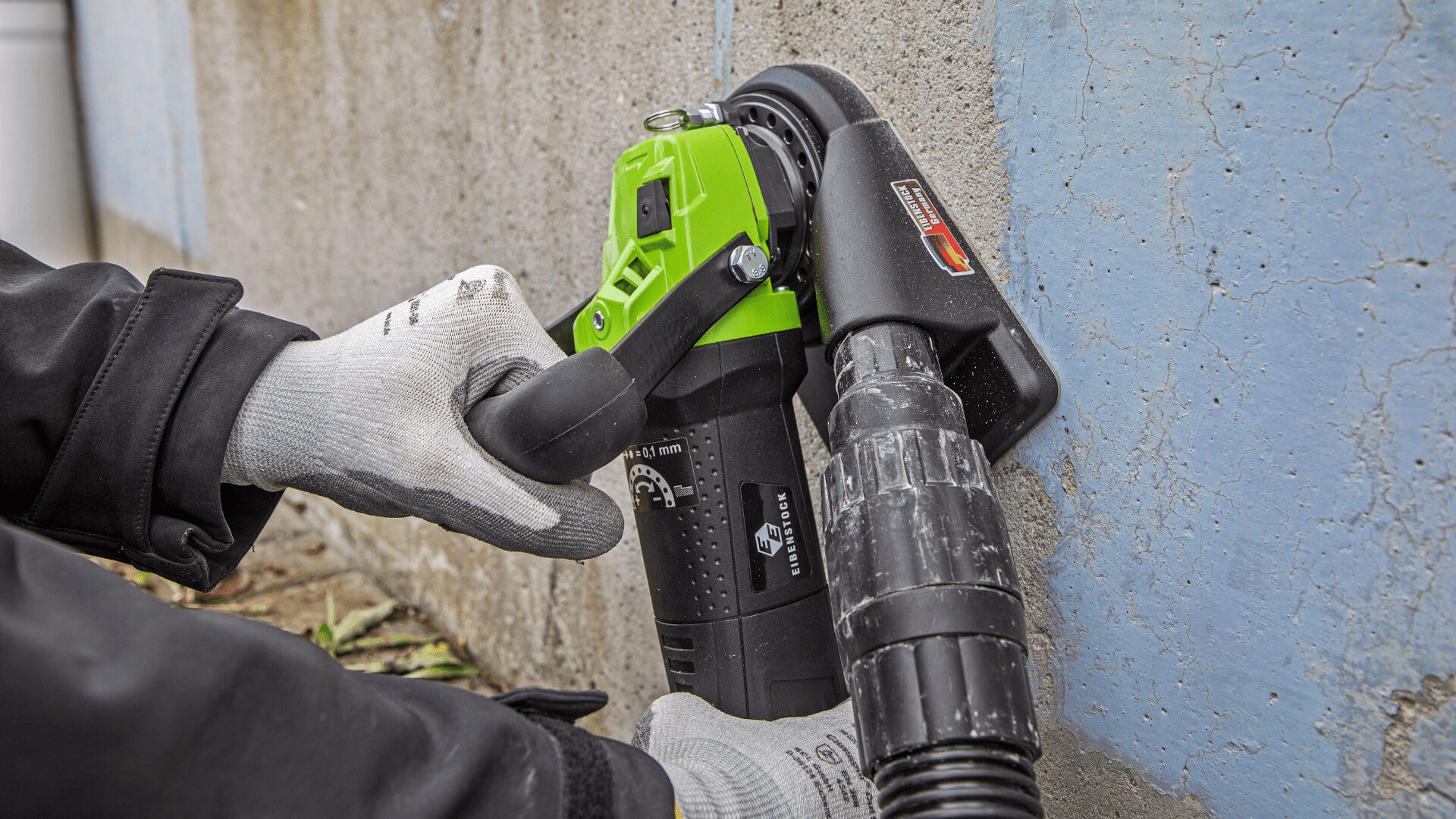 Eine Person mit Schutzhandschuhen schleift oder schleift mit einem grün-schwarzen Winkelschleifer mit Staubabsaugung eine Betonwand neben einem blau gestrichenen Abschnitt.