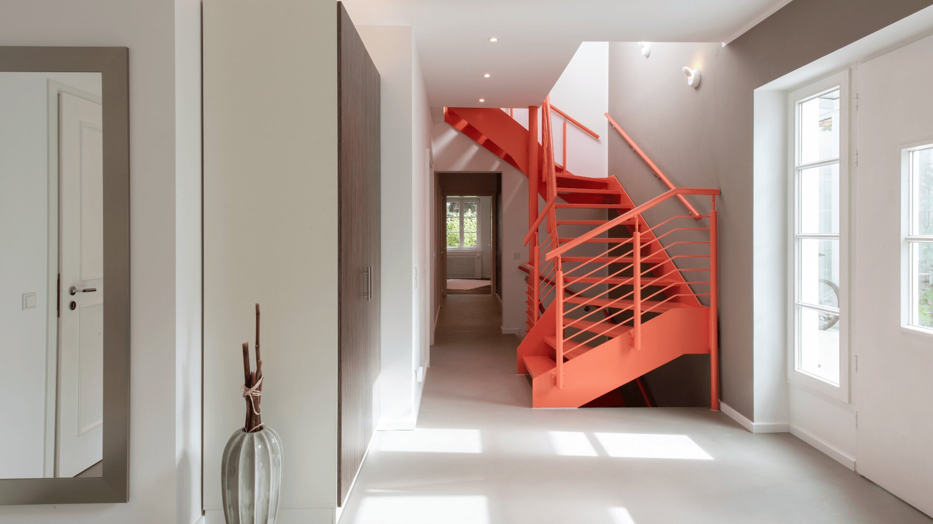 Ein heller Flur in einem modernen Haus verfügt über eine kräftige rote Wendeltreppe, große Fenster, die natürliches Licht hereinlassen, helle Böden und minimalistisches Dekor, darunter ein Spiegel und eine dekorative Vase.