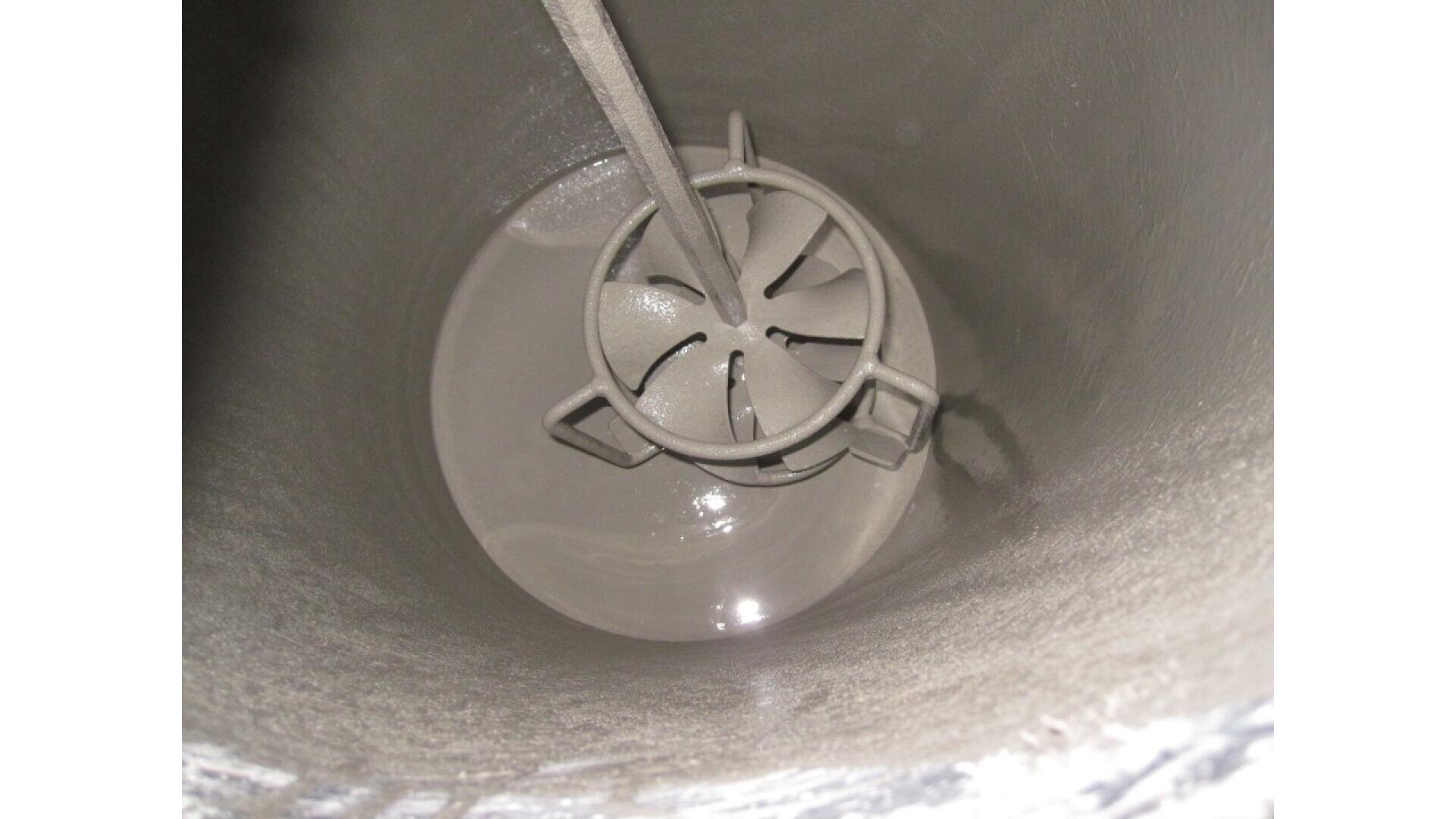 View looking down into a cylindrical tank, showing an industrial mixer with multiple angled blades attached to a central shaft, partly submerged in a thick, gray liquid.