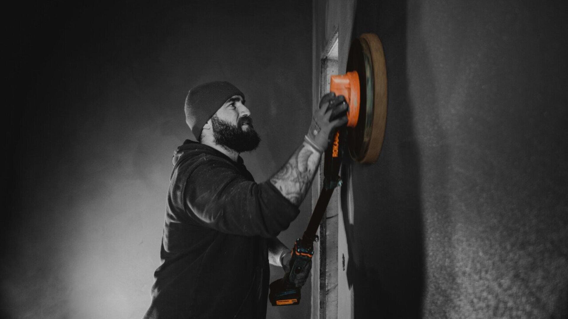 A bearded man wearing a beanie and hoodie uses a power sander on a wall, working intently in a dimly lit room with dramatic lighting.