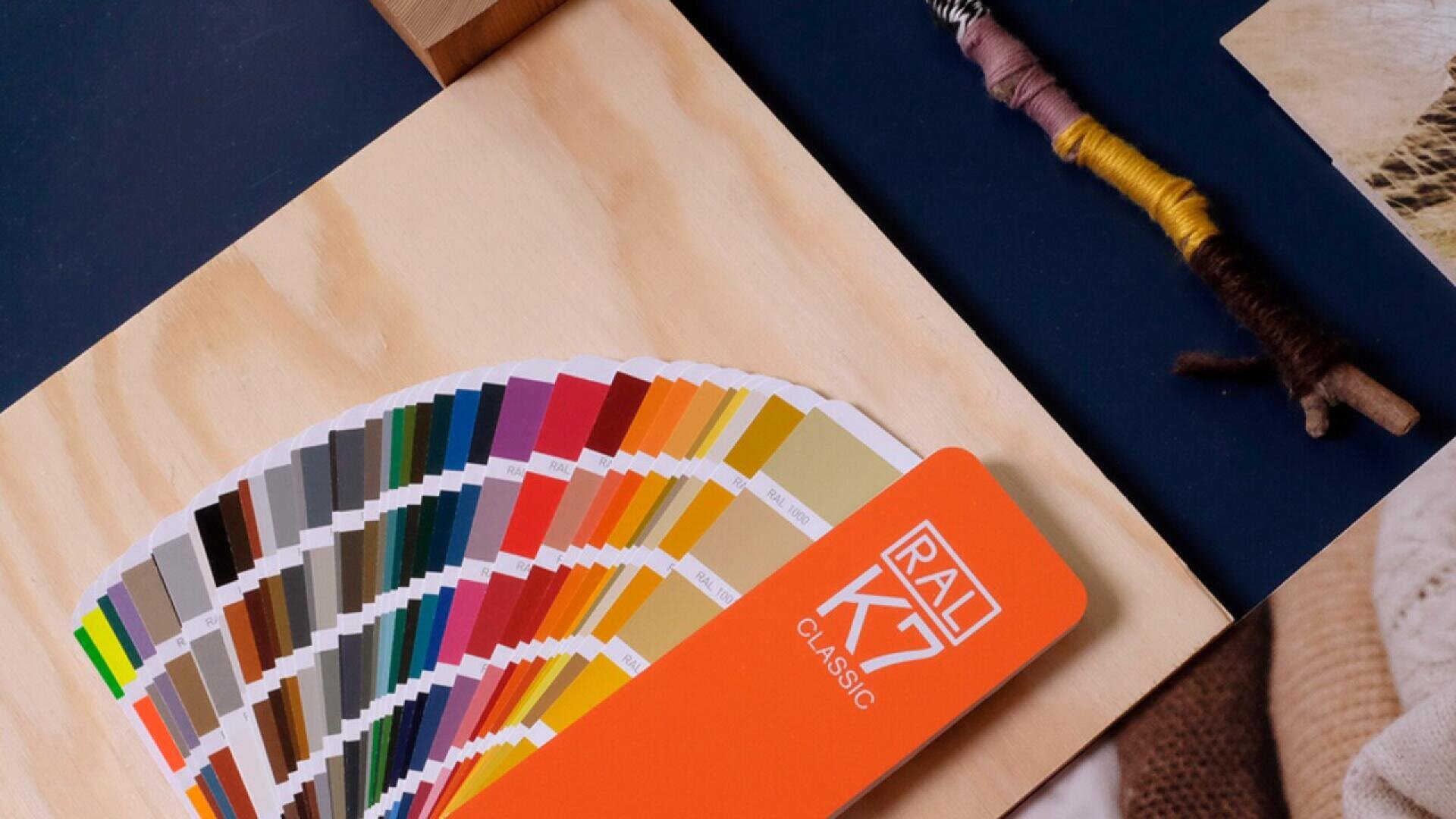 A RAL K7 Classic color fan deck is spread out on a wooden surface, displaying a variety of color swatches. Nearby are yarn-wrapped sticks and fabric, suggesting a creative or design workspace.