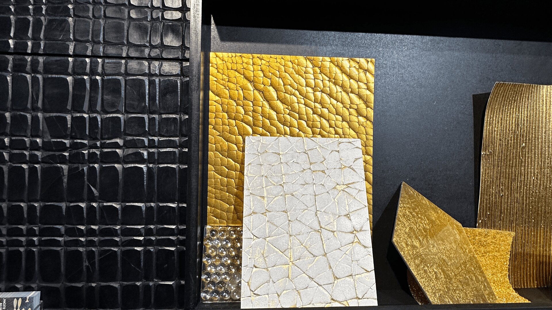 Eine Ausstellung strukturierter Materialproben, darunter ein schwarzes Krokodilmuster, metallisches Gold und weiß rissige Oberflächen sowie Goldblech, alle vertikal vor einem dunklen Hintergrund angeordnet.