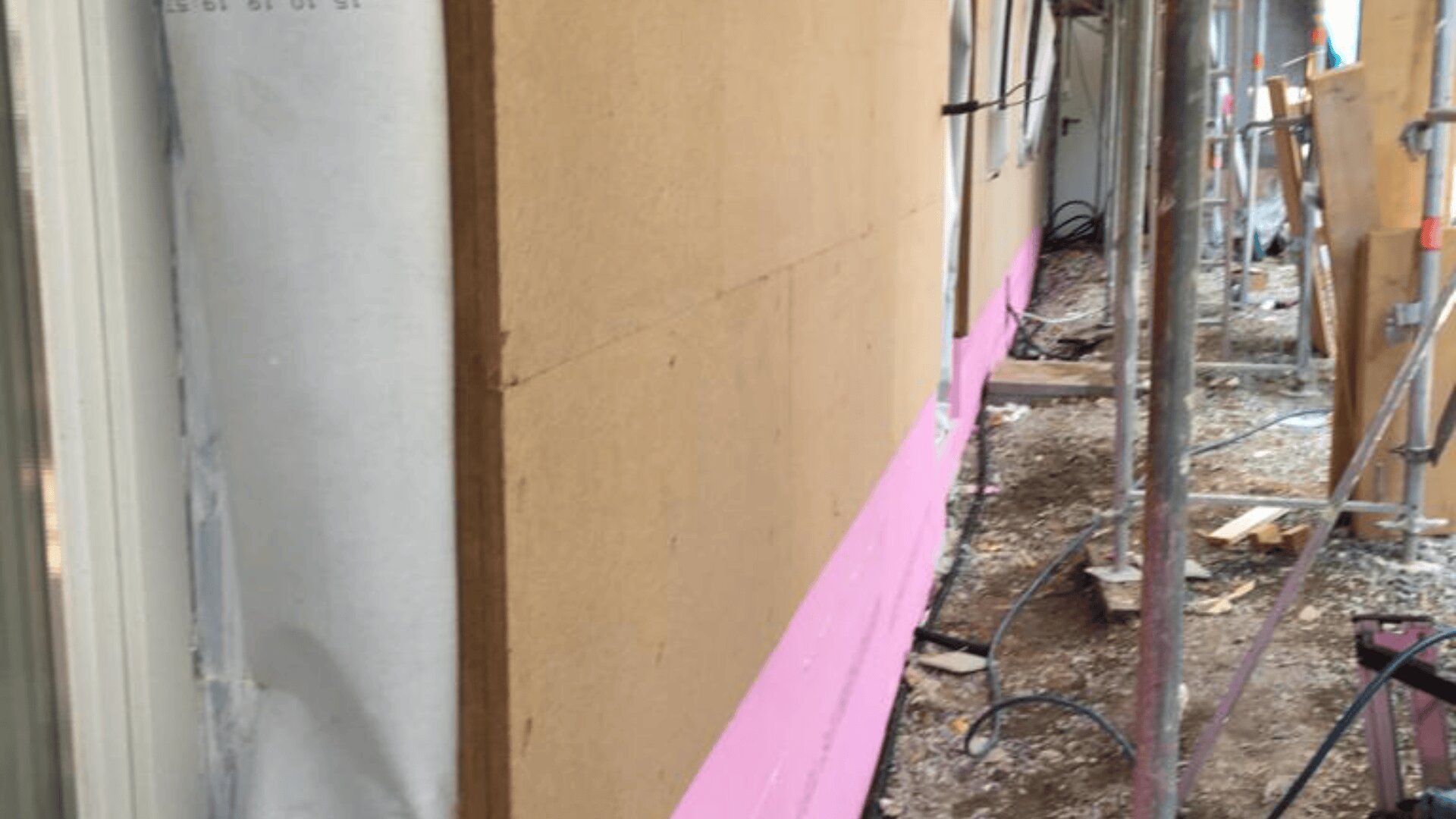 Ein im Bau befindliches Gebäude mit braunen Paneelen über einer weißen Wetterschutzwand und rosafarbener Isolierung am unteren Rand. Gerüste, Schutt und Stromkabel sind auf dem Boden neben der Wand sichtbar.